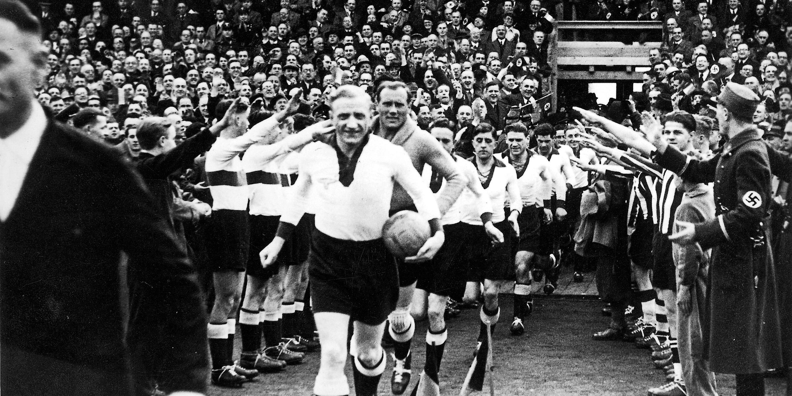 Die Ausstellung 'Fußball in der NS-Zeit' des Kreismuseums Wewelsburg kommt im Januar 2027 nach Saarbrücken. Historische Schwarz-Weiß-Fotografie eines Fußballstadions. Eine Reihe von Fußballspielern läuft aus einem Tunnel auf das Spielfeld. Die Person an der Spitze trägt einen Ball unter dem Arm und grüßt mit erhobener Hand. Links und rechts des Weges stehen zahlreiche Menschen dicht gedrängt, darunter auch uniformierte Personen; viele strecken den rechten Arm aus. Die Tribünen im Hintergrund sind bis auf den letzten Platz mit Zuschauern gefüllt. Die Aufnahme vermittelt den Eindruck eines großen Sportereignisses in einer politisch geprägten Atmosphäre der 1930er- oder 1940er-Jahre.
