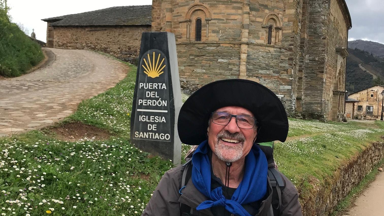 Ein älterer Mann mit Brille und großem Sonnenhut steht lächelnd vor einer alten romanischen Kirche aus Stein mit halbrunder Apsis. Neben ihm steht ein Wegweiser mit der Aufschrift „Puerta del Perdón – Iglesia de Santiago“, was auf den Jakobsweg hinweist. Im Hintergrund führen ein gepflasterter Weg und eine Wiese mit Gänseblümchen zur Kirche. (Alt-Text erstellt mit Unterstützung von ChatGPT)
