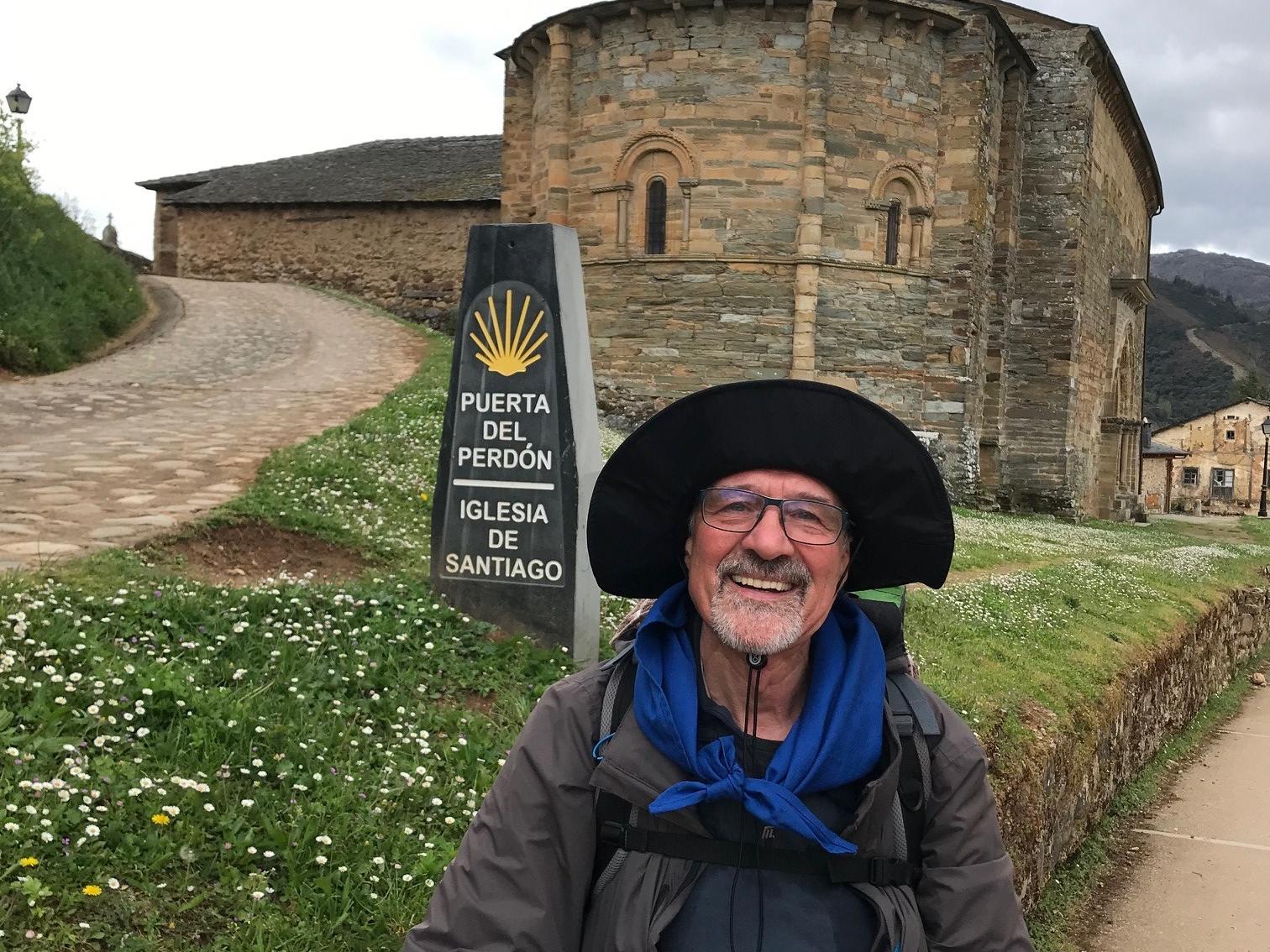 Ein älterer Mann mit Brille und großem Sonnenhut steht lächelnd vor einer alten romanischen Kirche aus Stein mit halbrunder Apsis. Neben ihm steht ein Wegweiser mit der Aufschrift „Puerta del Perdón – Iglesia de Santiago“, was auf den Jakobsweg hinweist. Im Hintergrund führen ein gepflasterter Weg und eine Wiese mit Gänseblümchen zur Kirche. (Alt-Text erstellt mit Unterstützung von ChatGPT)