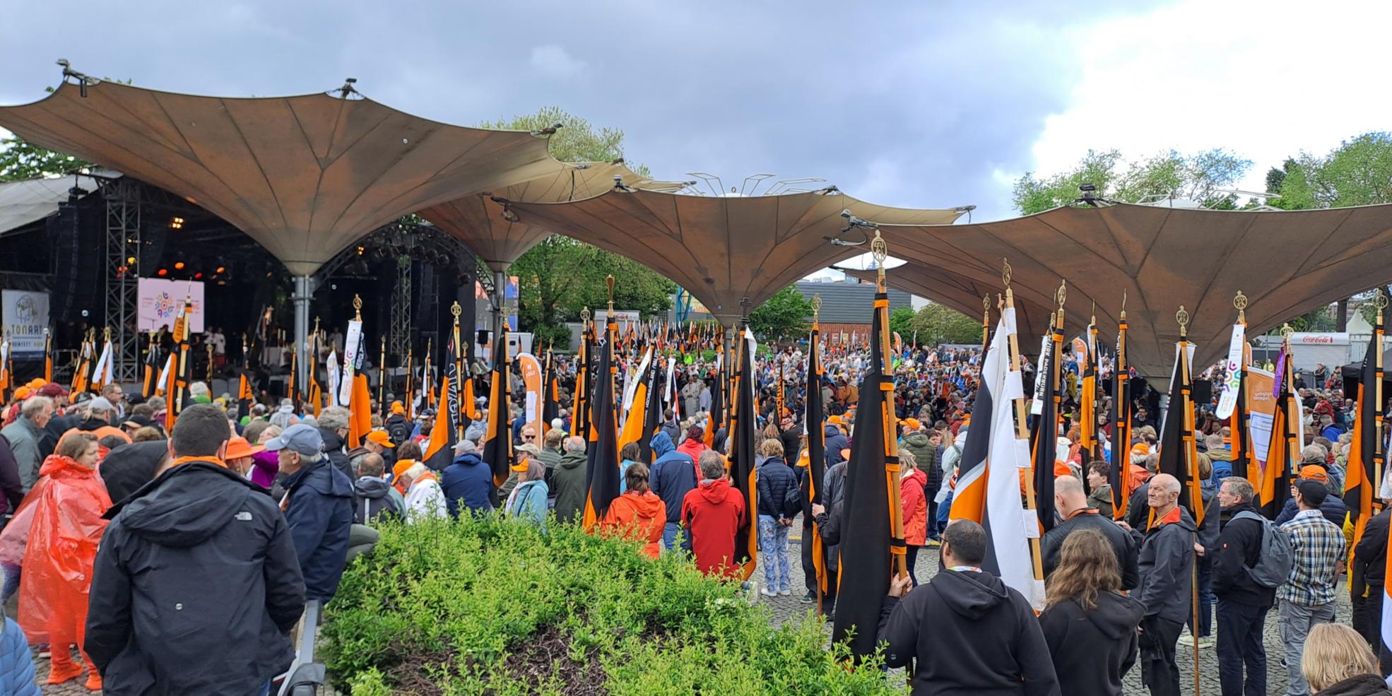 Auf dem Bild ist eine große Menschenmenge bei einer Veranstaltung im Freien zu sehen, viele von ihnen tragen orangefarbene Kleidung oder Accessoires. Zahlreiche orange-schwarze Fahnen mit goldenen Spitzen werden aufrecht gehalten. Im Hintergrund befindet sich eine überdachte Bühne mit Lautsprechern und Bildschirmen unter großen, zeltartigen Dächern. (Alt-Text erstellt mit Unterstützung von ChatGPT)