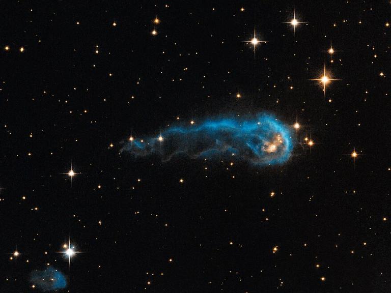 Das Weltraum-Foto zeigt das schwarze Weltall mit weißlich bis gelblich leuchtenden Sternen von etwa einem bis fünf Millimetern Größe. In der Bildmitte eine längliche, außen blau leuchtende Wolke.