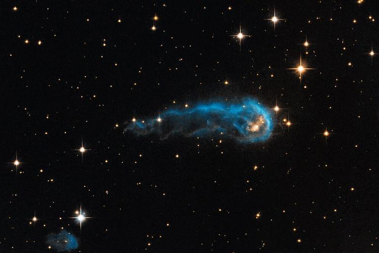 Das Weltraum-Foto zeigt das schwarze Weltall mit weißlich bis gelblich leuchtenden Sternen von etwa einem bis fünf Millimetern Größe. In der Bildmitte eine längliche, außen blau leuchtende Wolke.