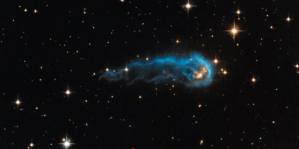 Das Weltraum-Foto zeigt das schwarze Weltall mit weißlich bis gelblich leuchtenden Sternen von etwa einem bis fünf Millimetern Größe. In der Bildmitte eine längliche, außen blau leuchtende Wolke.