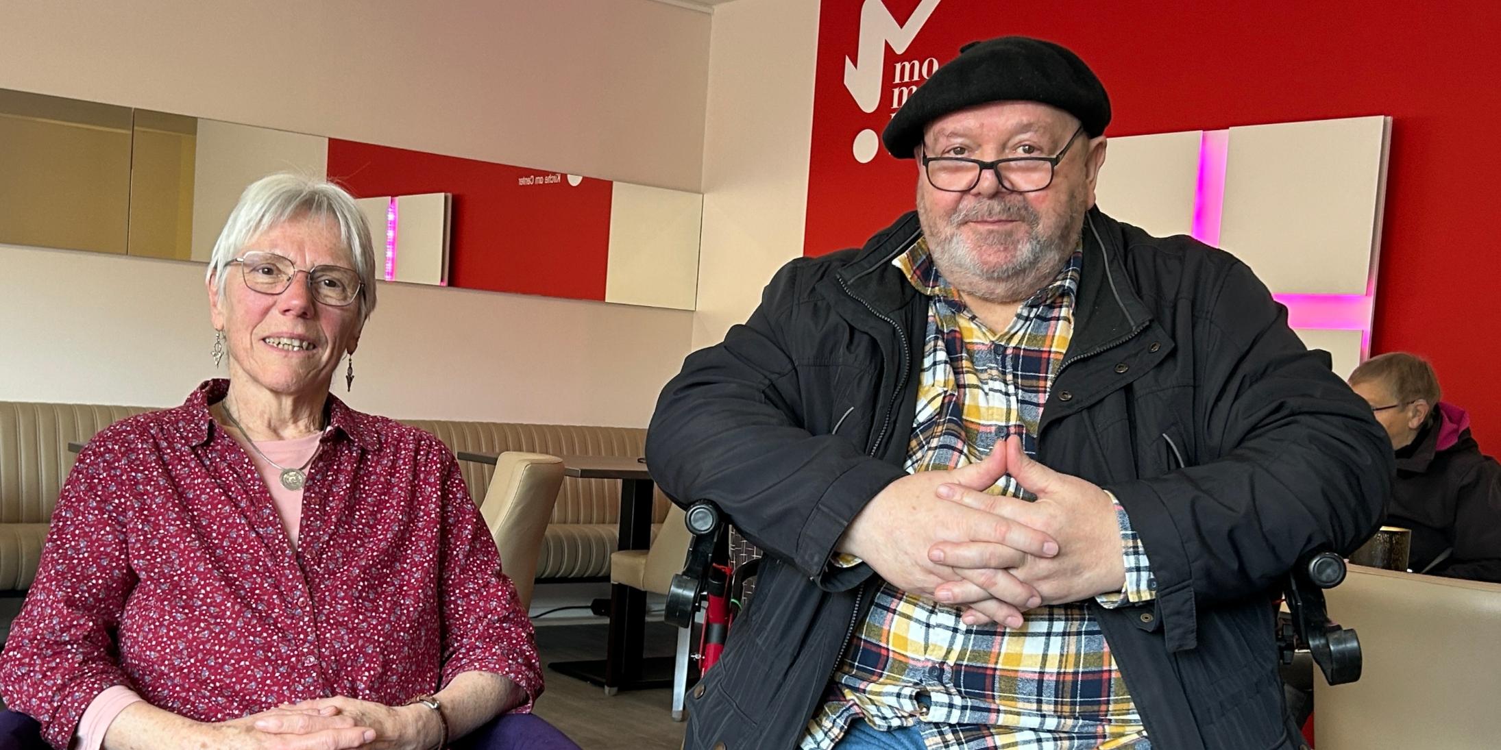 Eine Frau mit Brille sitzt neben einem Mann, der ebenfalls eine Brille und eine Mütze trägt. Beide schauen in die Kamera