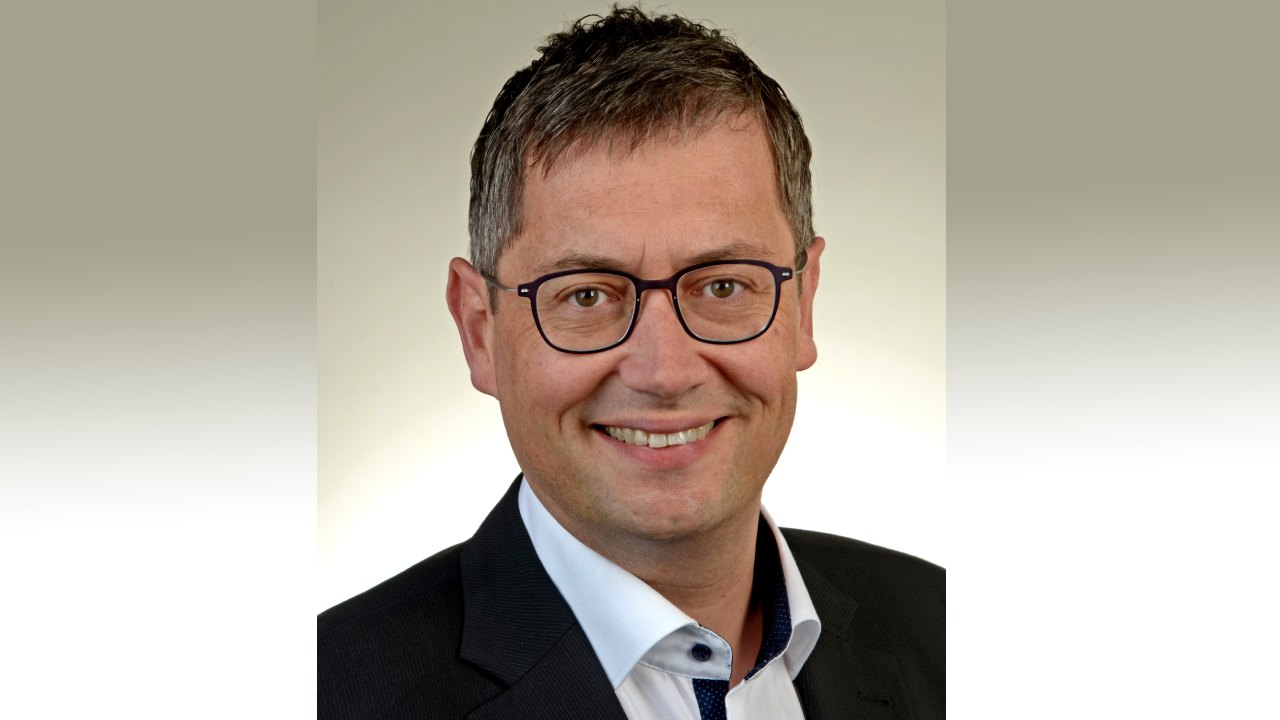 Ein Mann, mittleren Alters, mit Brille und kurzen Haaren, lächelt in die Kamera. Er trägt einen schwarzen Anzug ohne Krawatte
