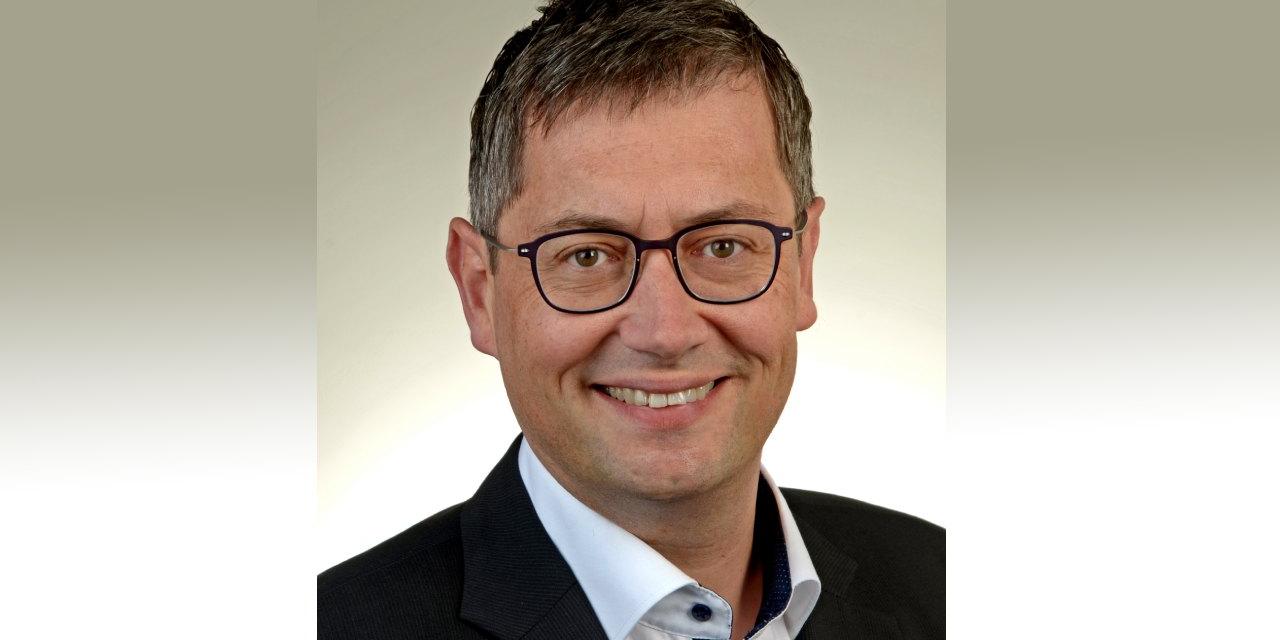 Ein Mann, mittleren Alters, mit Brille und kurzen Haaren, lächelt in die Kamera. Er trägt einen schwarzen Anzug ohne Krawatte