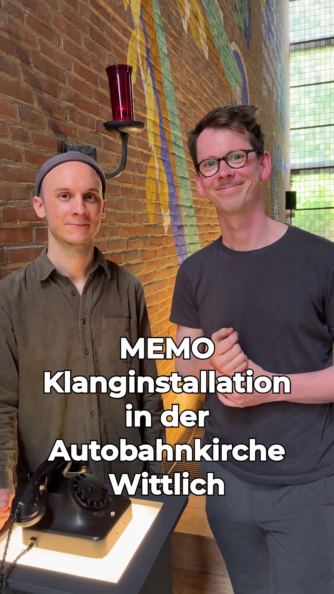 Man sieht zwei Männer, die vor einer bemalten und angestrahlten Backsteinmauer in der Kirche stehen