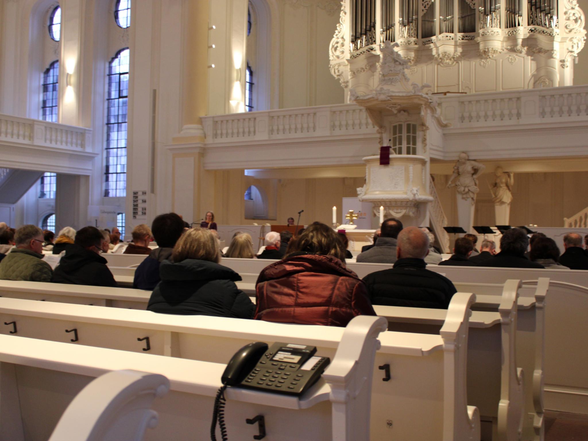 Menschen sitzen in den Kirchenbänken, auf den Bänken stehen Telefone