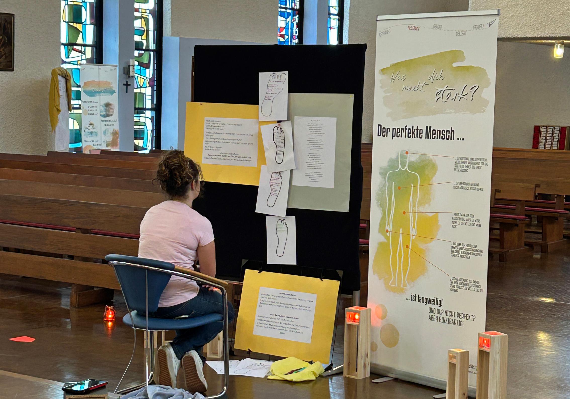 Eine junge Frau sitzt vor einer Plakatwand. Was macht dich stark wird auf einem großen Plakat gefragt.
