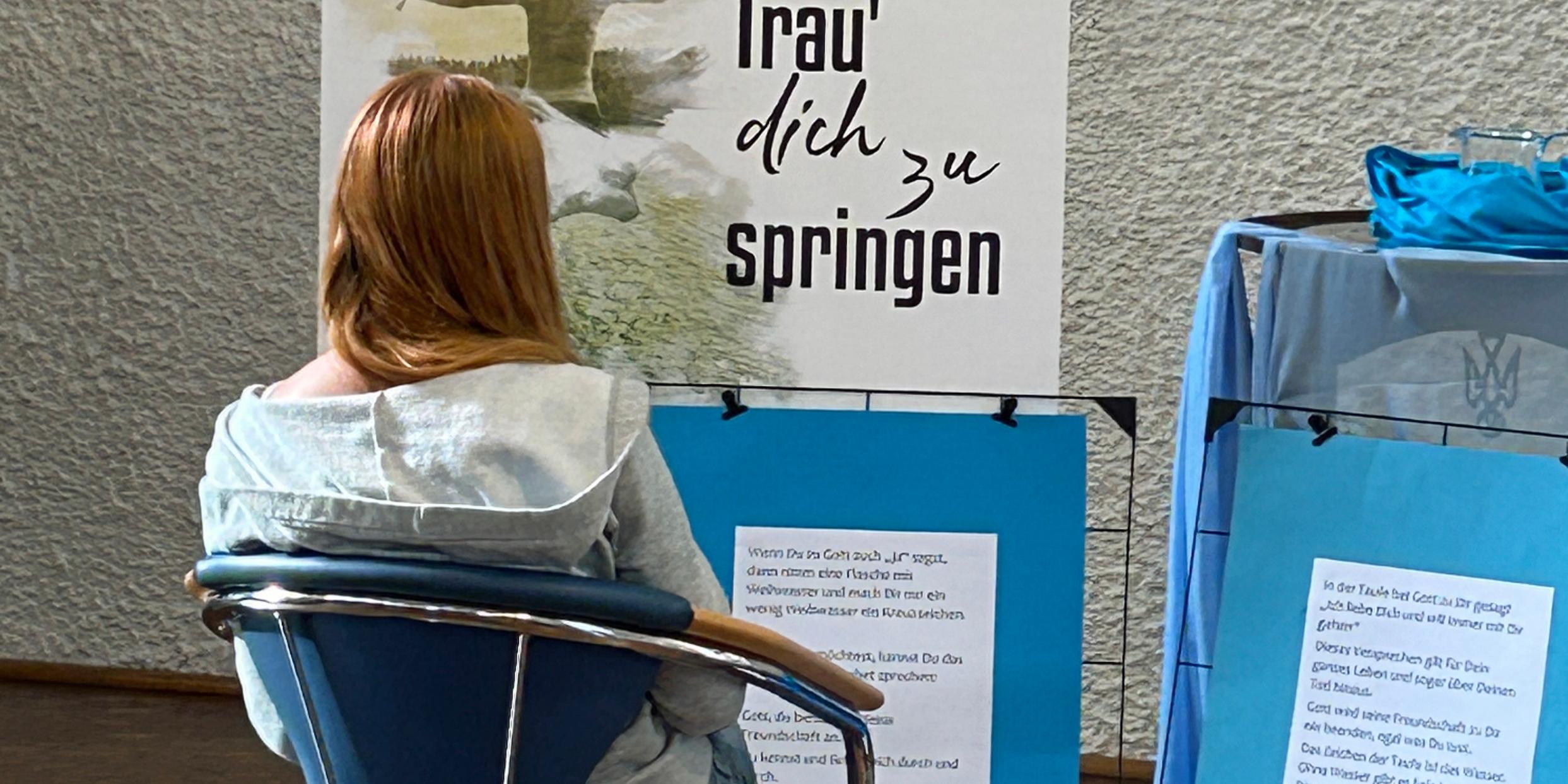Eine junge Frau sitzt auf einem Stuhl, vor ihr stehen Plakate mit der Aufschrift Trau dich zu springen und weiteren Texten.
