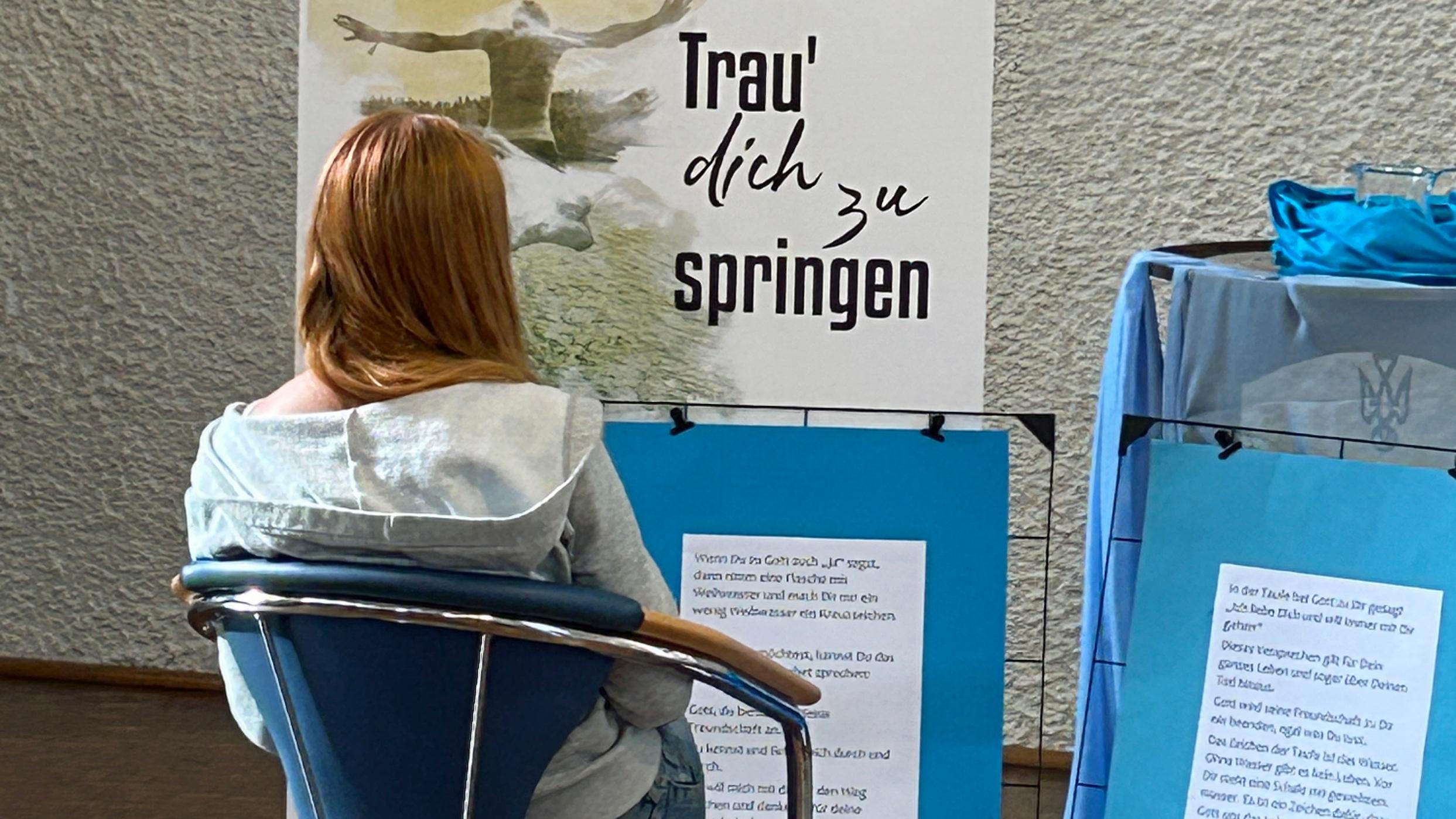 Eine junge Frau sitzt auf einem Stuhl, vor ihr stehen Plakate mit der Aufschrift Trau dich zu springen und weiteren Texten.