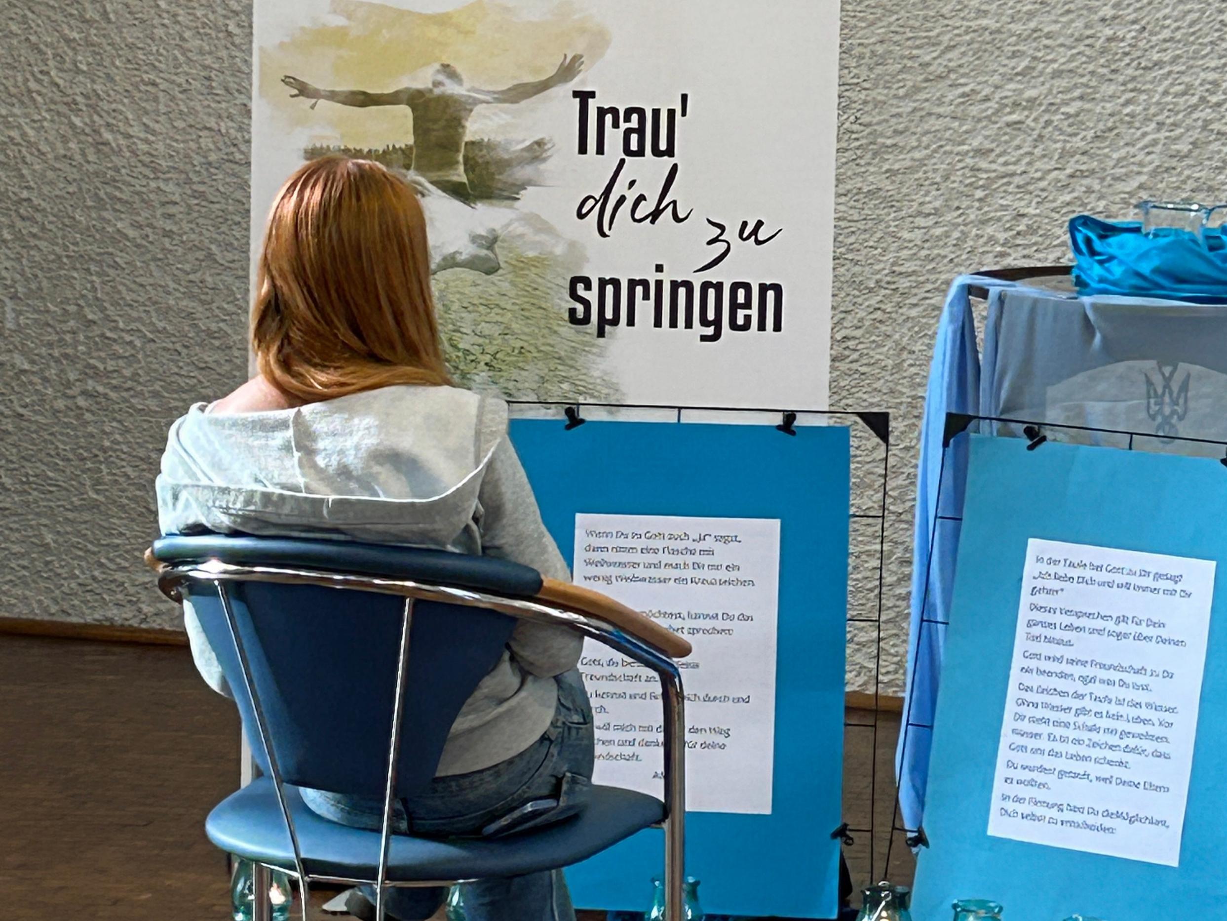 Eine junge Frau sitzt auf einem Stuhl, vor ihr stehen Plakate mit der Aufschrift Trau dich zu springen und weiteren Texten.