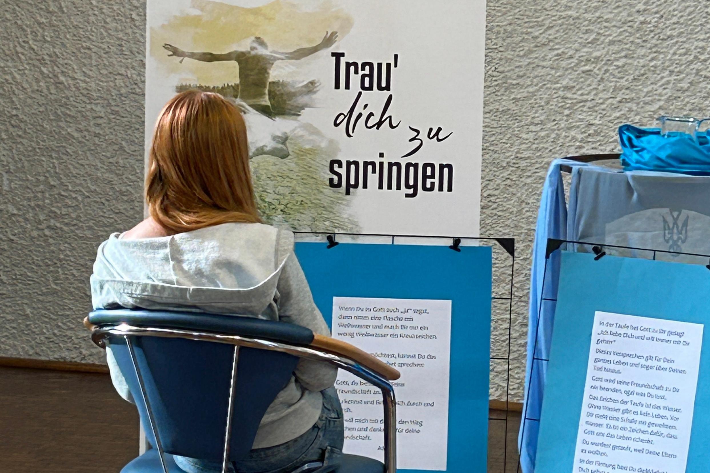 Eine junge Frau sitzt auf einem Stuhl, vor ihr stehen Plakate mit der Aufschrift Trau dich zu springen und weiteren Texten.