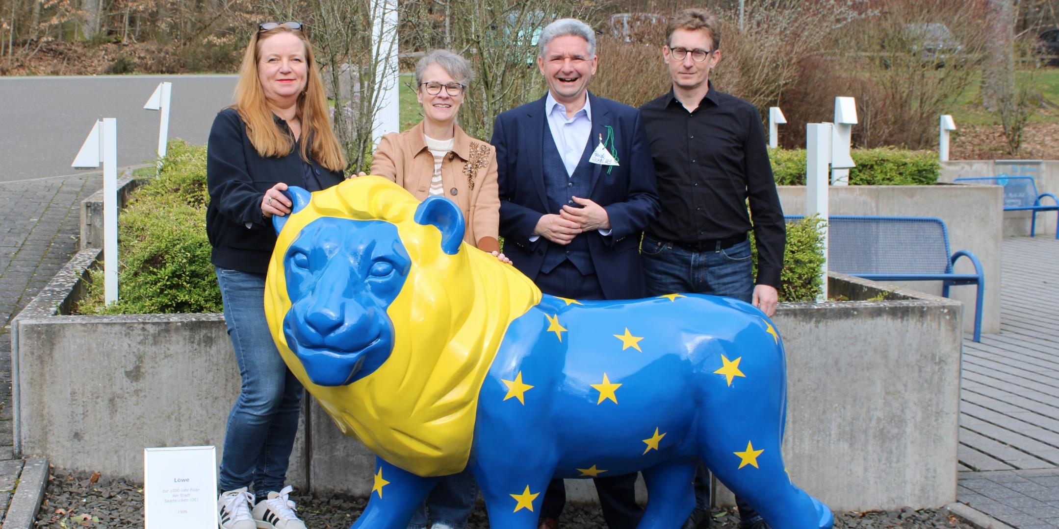 Vier Personen stehen im Freien hinter einer großen, bunt bemalten Löwenfigur. Der Löwe ist in kräftigem Blau und Gelb gestaltet und trägt gelbe Sterne auf dem Körper. Die Personen stehen dicht beieinander, einige berühren die Figur. Rechts und links befinden sich Betonpflanzkübel, dahinter sind Büsche, Wege und ein paar Sitzbänke zu sehen. Ein kleines Schild steht links neben der Figur. Geben Sie Ihr Feedback zu BizChat ab.