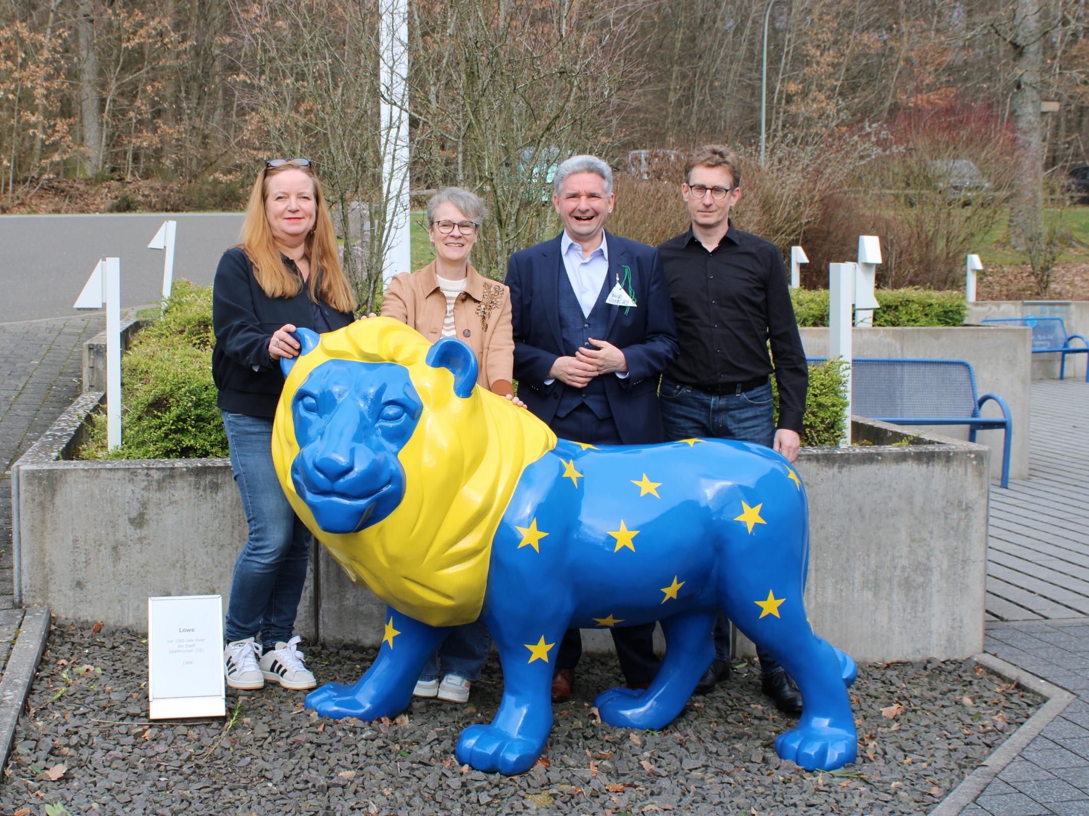 Vier Personen stehen im Freien hinter einer großen, bunt bemalten Löwenfigur. Der Löwe ist in kräftigem Blau und Gelb gestaltet und trägt gelbe Sterne auf dem Körper. Die Personen stehen dicht beieinander, einige berühren die Figur. Rechts und links befinden sich Betonpflanzkübel, dahinter sind Büsche, Wege und ein paar Sitzbänke zu sehen. Ein kleines Schild steht links neben der Figur. Geben Sie Ihr Feedback zu BizChat ab.