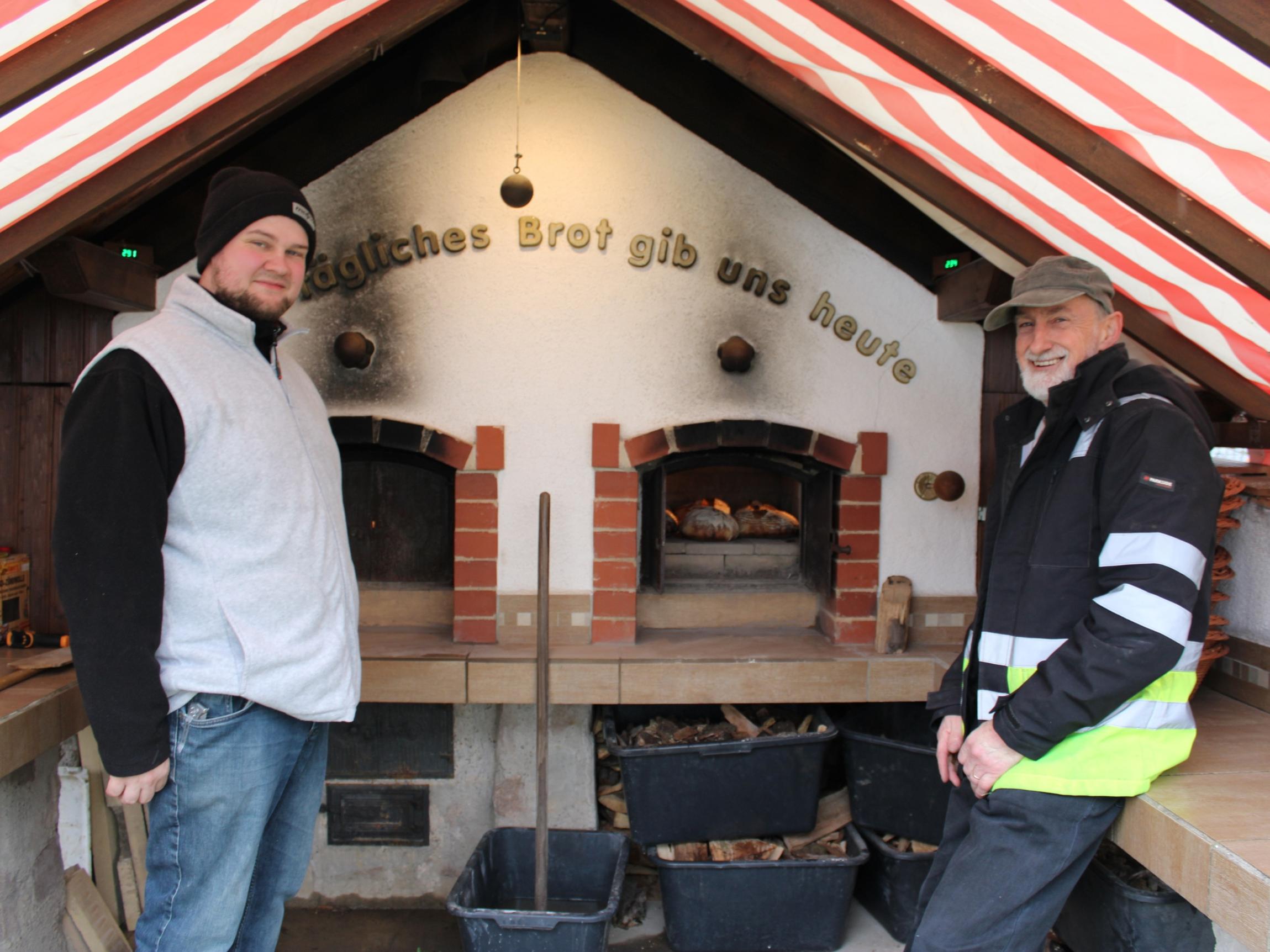 In einem überdachten Außenbereich stehen zwei Personen vor einem großen, gemauerten Holzbackofen. Der Ofen besitzt zwei Backkammern, in denen Brotlaibe auf Steinflächen liegen und backen. Über dem Ofen ist der Schriftzug „Tägliches Brot gib uns heute“ angebracht. Unterhalb der Arbeitsfläche stehen mehrere schwarze Kunststoffwannen mit Holzstücken. Das Dach besteht aus schräg verlaufenden Balken und ist mit einer rot-weiß gestreiften Plane überspannt.