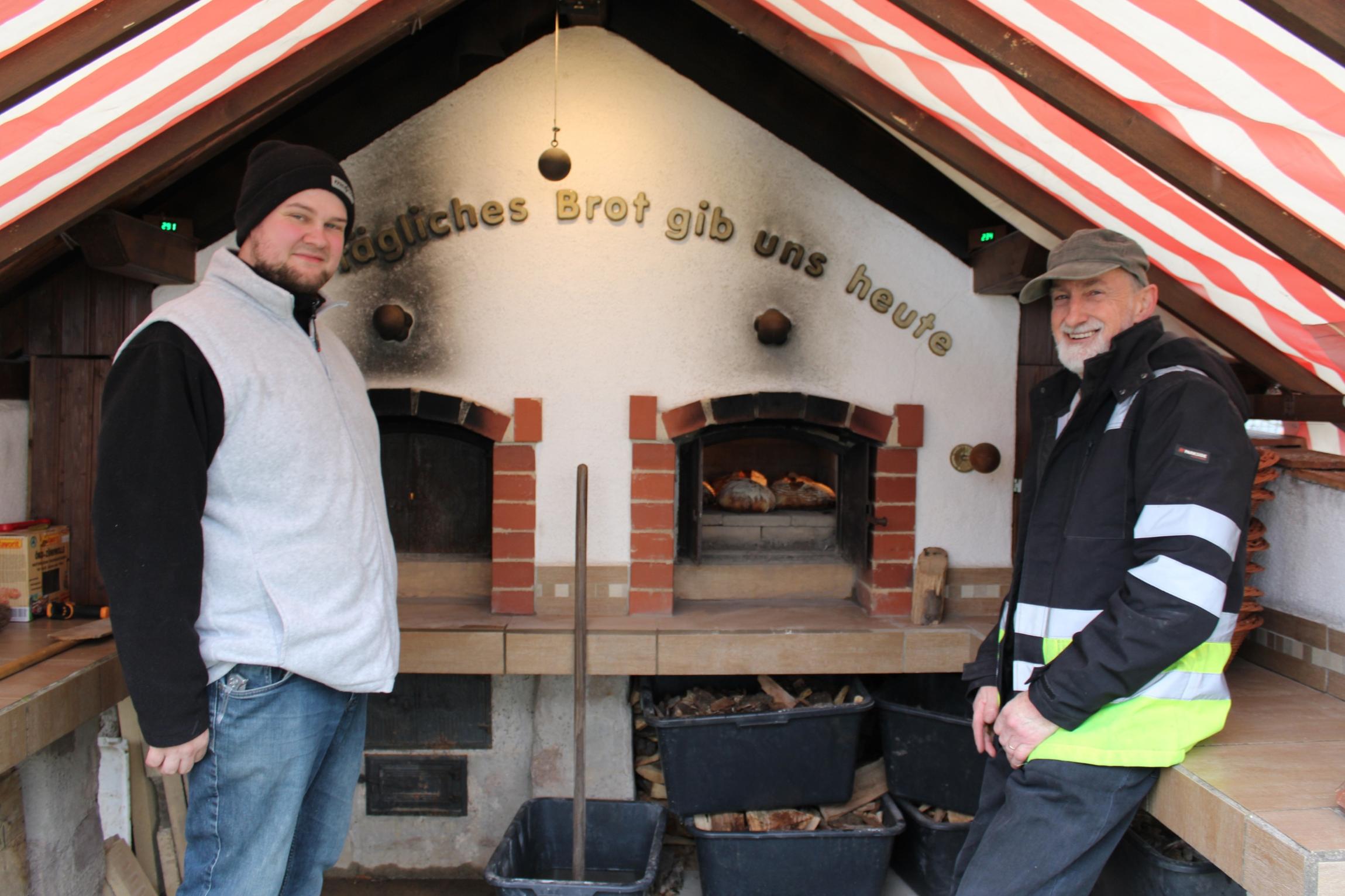In einem überdachten Außenbereich stehen zwei Personen vor einem großen, gemauerten Holzbackofen. Der Ofen besitzt zwei Backkammern, in denen Brotlaibe auf Steinflächen liegen und backen. Über dem Ofen ist der Schriftzug „Tägliches Brot gib uns heute“ angebracht. Unterhalb der Arbeitsfläche stehen mehrere schwarze Kunststoffwannen mit Holzstücken. Das Dach besteht aus schräg verlaufenden Balken und ist mit einer rot-weiß gestreiften Plane überspannt.