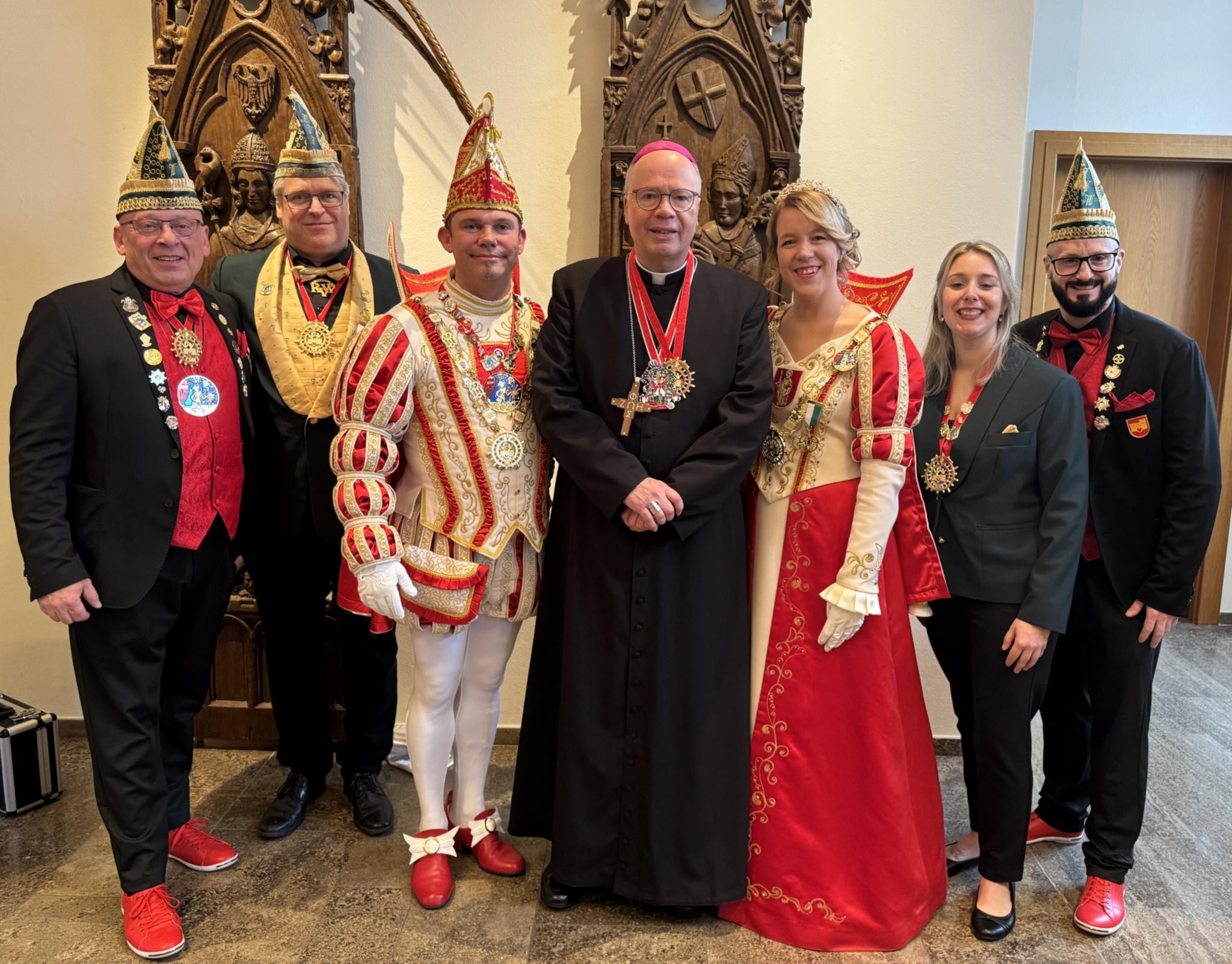 Bischof Ackermann in schwarzer liturgischer Kleidung steht in der Mitte einer festlich gekleideten Gruppe. Neben ihm posieren ein Prinz in rot‑weißem Karnevalskostüm und eine Prinzessin in einem rot‑goldenen Kleid. Weitere Personen tragen Anzüge, Karnevalsorden und bunte Narrenkappen