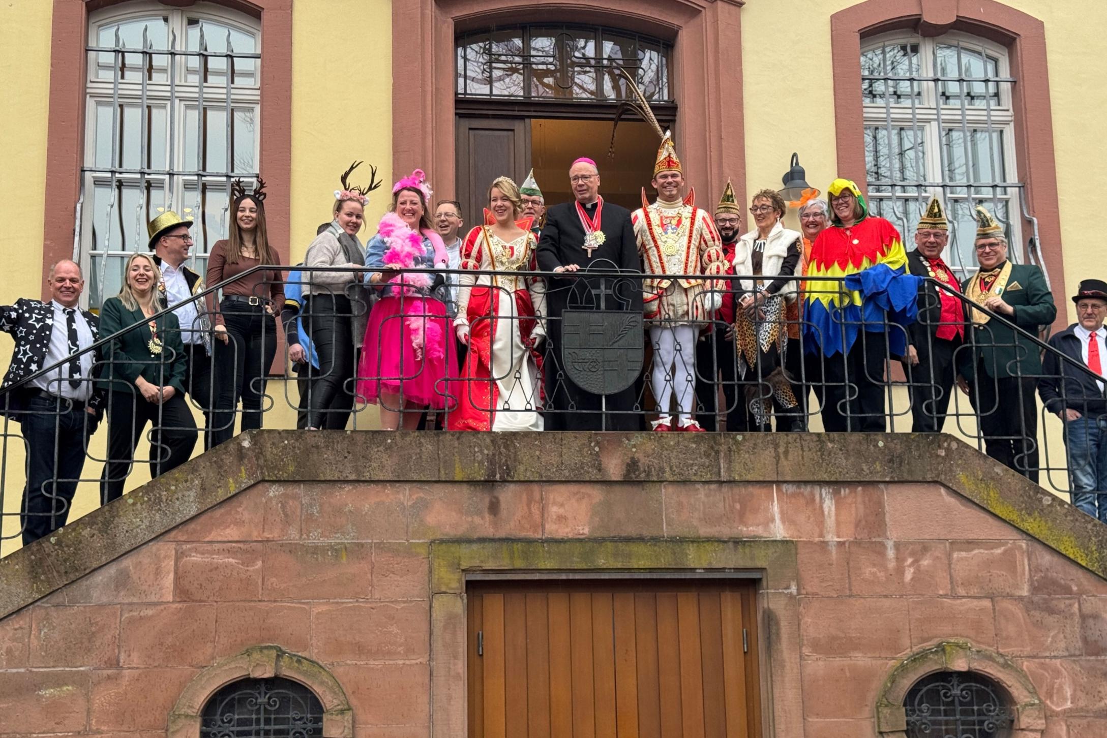 Eine große Gruppe steht auf einem Balkongang vor einem historischen Gebäude mit hellen Wänden und hohen Fenstern. In der Mitte befindet sich Bischof Ackermann in dunkler liturgischer Kleidung. Neben ihm stehen das Prinzenpaar in aufwendigen rot‑weißen und rot‑goldenen Karnevalskostümen sowie Angehörige ihres Gefolges mit traditionellen Narrenkappen. Rundherum stehen Mitarbeitende des Bischofshofes und des Generalvikariates.