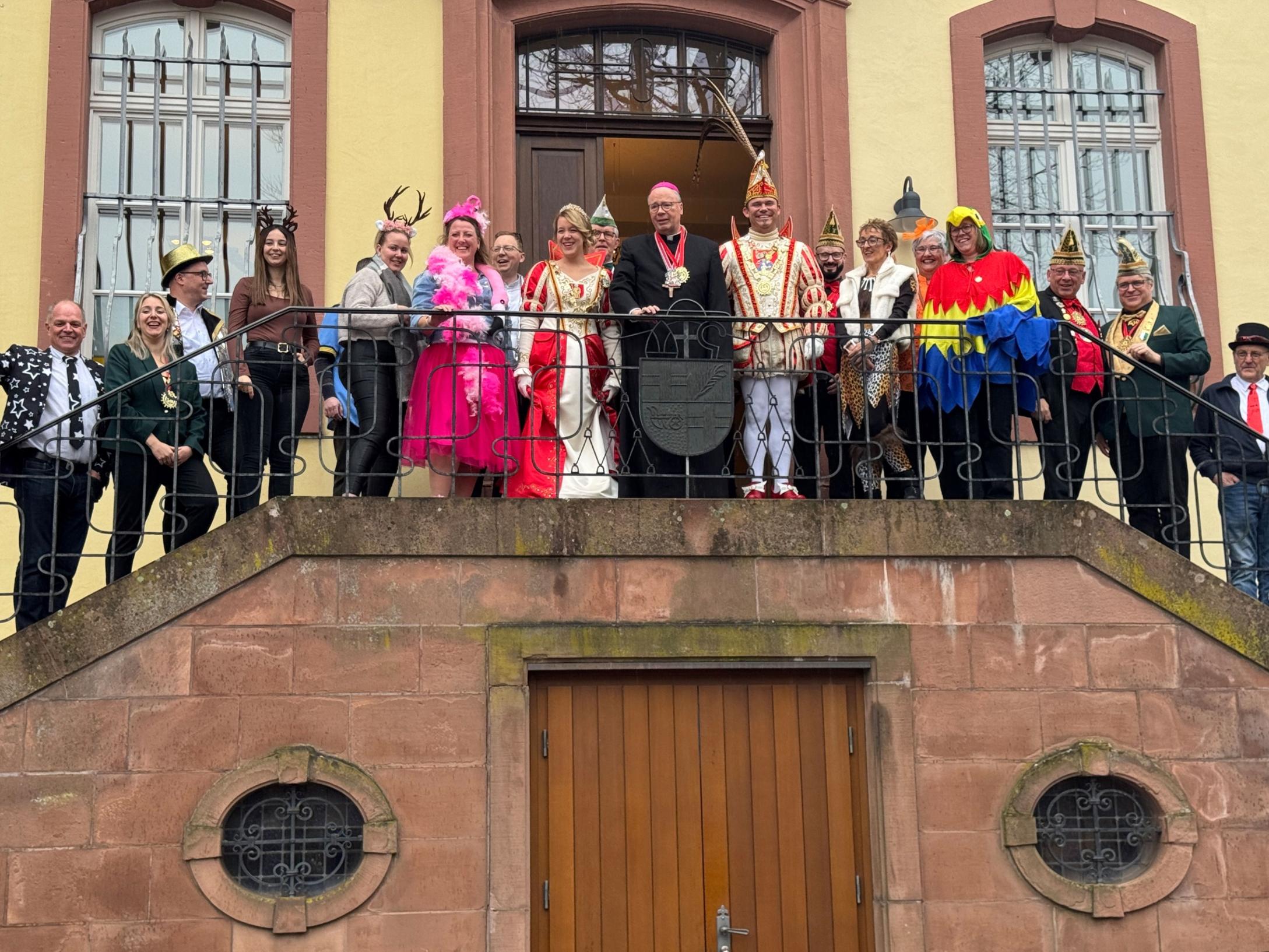 Eine große Gruppe steht auf einem Balkongang vor einem historischen Gebäude mit hellen Wänden und hohen Fenstern. In der Mitte befindet sich Bischof Ackermann in dunkler liturgischer Kleidung. Neben ihm stehen das Prinzenpaar in aufwendigen rot‑weißen und rot‑goldenen Karnevalskostümen sowie Angehörige ihres Gefolges mit traditionellen Narrenkappen. Rundherum stehen Mitarbeitende des Bischofshofes und des Generalvikariates.