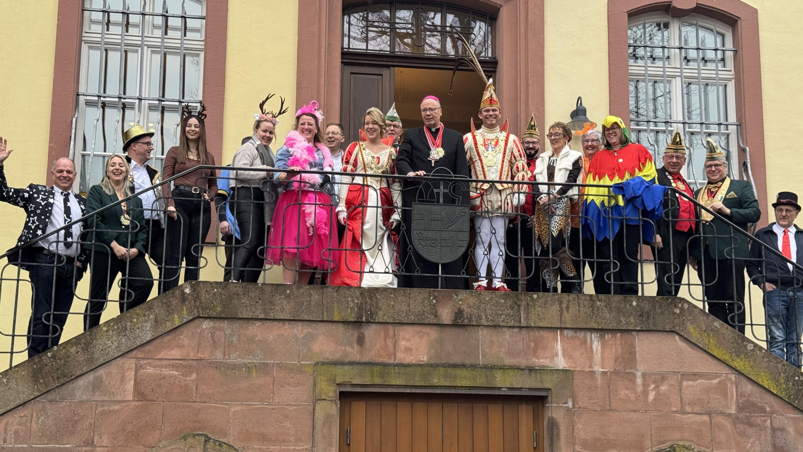 Eine große Gruppe steht auf einem Balkongang vor einem historischen Gebäude mit hellen Wänden und hohen Fenstern. In der Mitte befindet sich Bischof Ackermann in dunkler liturgischer Kleidung. Neben ihm stehen das Prinzenpaar in aufwendigen rot‑weißen und rot‑goldenen Karnevalskostümen sowie Angehörige ihres Gefolges mit traditionellen Narrenkappen. Rundherum stehen Mitarbeitende des Bischofshofes und des Generalvikariates.