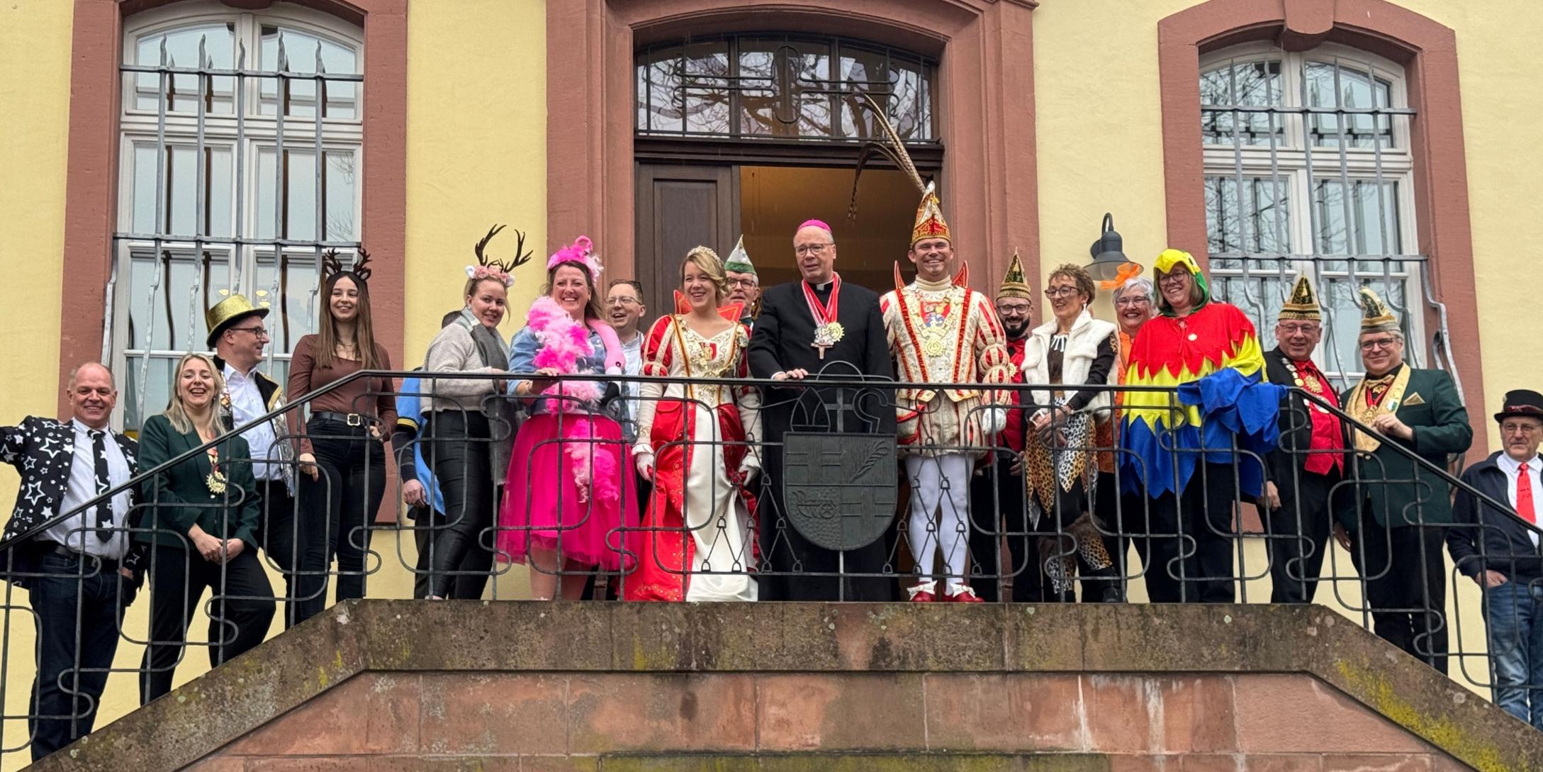 Eine große Gruppe steht auf einem Balkongang vor einem historischen Gebäude mit hellen Wänden und hohen Fenstern. In der Mitte befindet sich Bischof Ackermann in dunkler liturgischer Kleidung. Neben ihm stehen das Prinzenpaar in aufwendigen rot‑weißen und rot‑goldenen Karnevalskostümen sowie Angehörige ihres Gefolges mit traditionellen Narrenkappen. Rundherum stehen Mitarbeitende des Bischofshofes und des Generalvikariates.