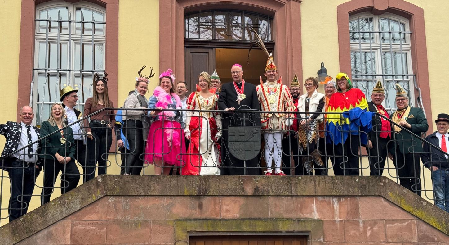 Eine große Gruppe steht auf einem Balkongang vor einem historischen Gebäude mit hellen Wänden und hohen Fenstern. In der Mitte befindet sich Bischof Ackermann in dunkler liturgischer Kleidung. Neben ihm stehen das Prinzenpaar in aufwendigen rot‑weißen und rot‑goldenen Karnevalskostümen sowie Angehörige ihres Gefolges mit traditionellen Narrenkappen. Rundherum stehen Mitarbeitende des Bischofshofes und des Generalvikariates.