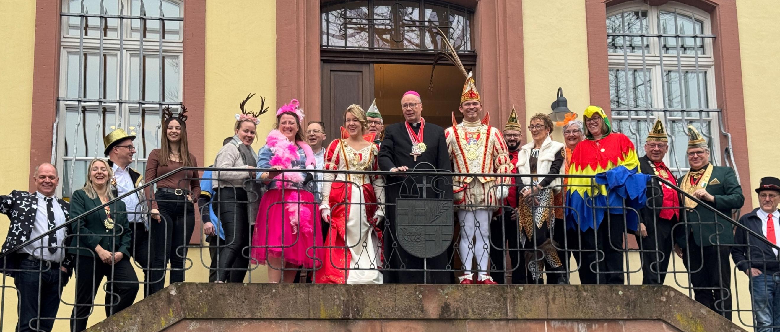 Eine große Gruppe steht auf einem Balkongang vor einem historischen Gebäude mit hellen Wänden und hohen Fenstern. In der Mitte befindet sich Bischof Ackermann in dunkler liturgischer Kleidung. Neben ihm stehen das Prinzenpaar in aufwendigen rot‑weißen und rot‑goldenen Karnevalskostümen sowie Angehörige ihres Gefolges mit traditionellen Narrenkappen. Rundherum stehen Mitarbeitende des Bischofshofes und des Generalvikariates.