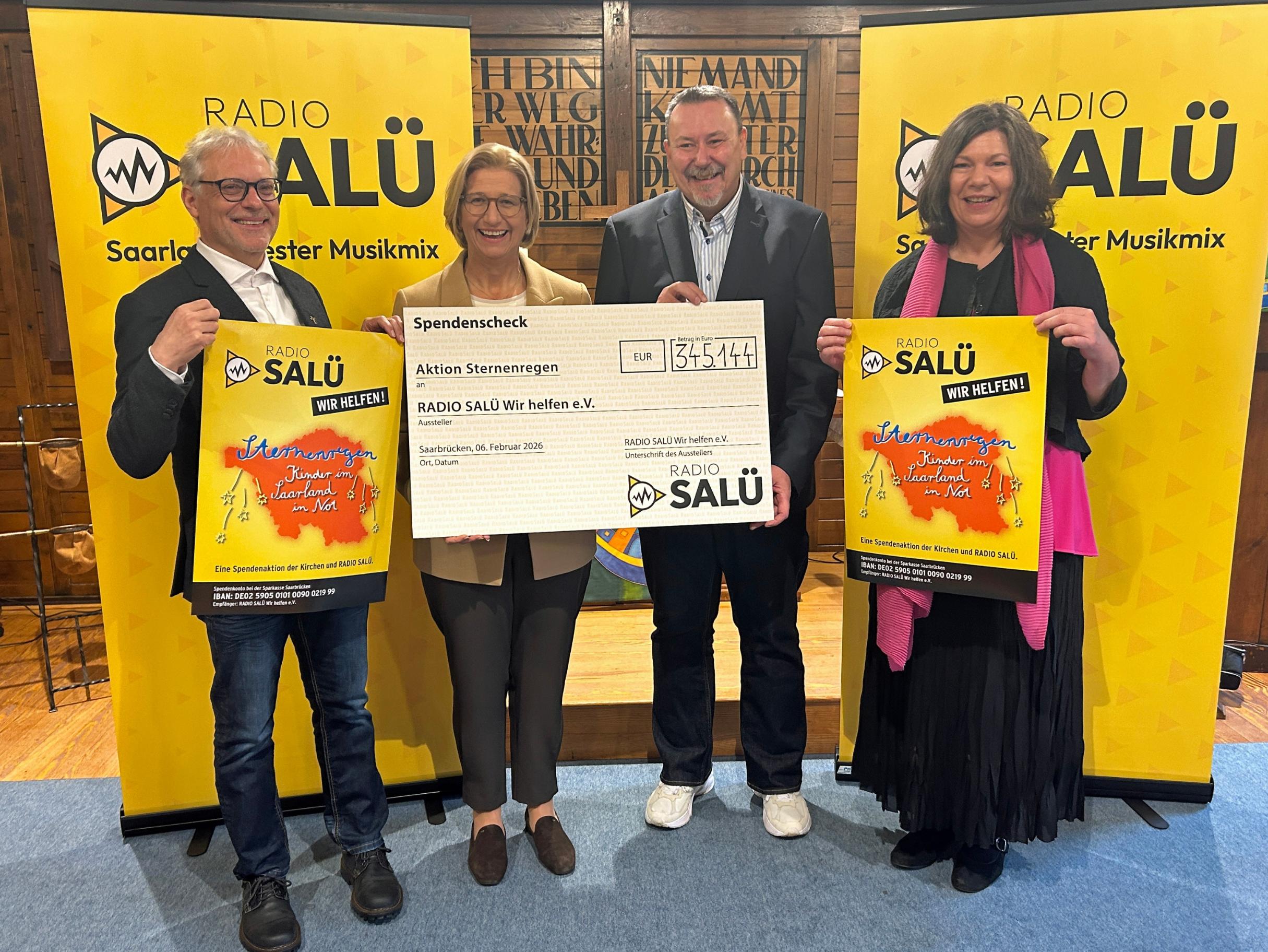 Vier Personen stehen in einem Innenraum vor zwei gelben Roll‑Up‑Bannern mit dem Logo von „Radio Salü“. Die beiden Personen in der Mitte halten gemeinsam einen großen Spendenscheck mit der Aufschrift „Aktion Sternenregen“ .Die beiden Personen außen halten jeweils ein farbiges Plakat derselben Spendenaktion. Der Boden besteht aus Holz, und im Hintergrund sind dunkle Holzwände mit eingravierten Schriftzügen zu sehen.