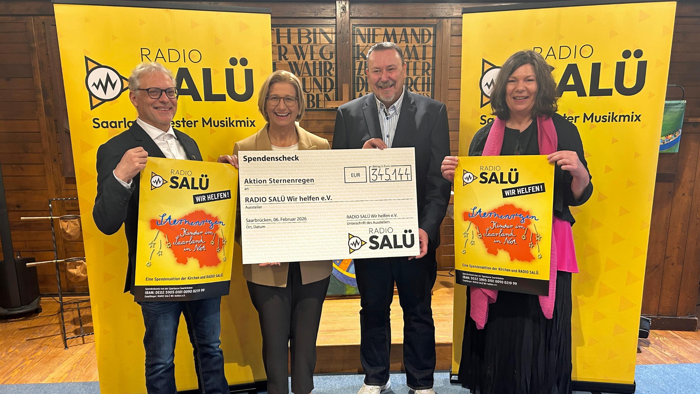 Vier Personen stehen in einem Innenraum vor zwei gelben Roll‑Up‑Bannern mit dem Logo von „Radio Salü“. Die beiden Personen in der Mitte halten gemeinsam einen großen Spendenscheck mit der Aufschrift „Aktion Sternenregen“ .Die beiden Personen außen halten jeweils ein farbiges Plakat derselben Spendenaktion. Der Boden besteht aus Holz, und im Hintergrund sind dunkle Holzwände mit eingravierten Schriftzügen zu sehen.