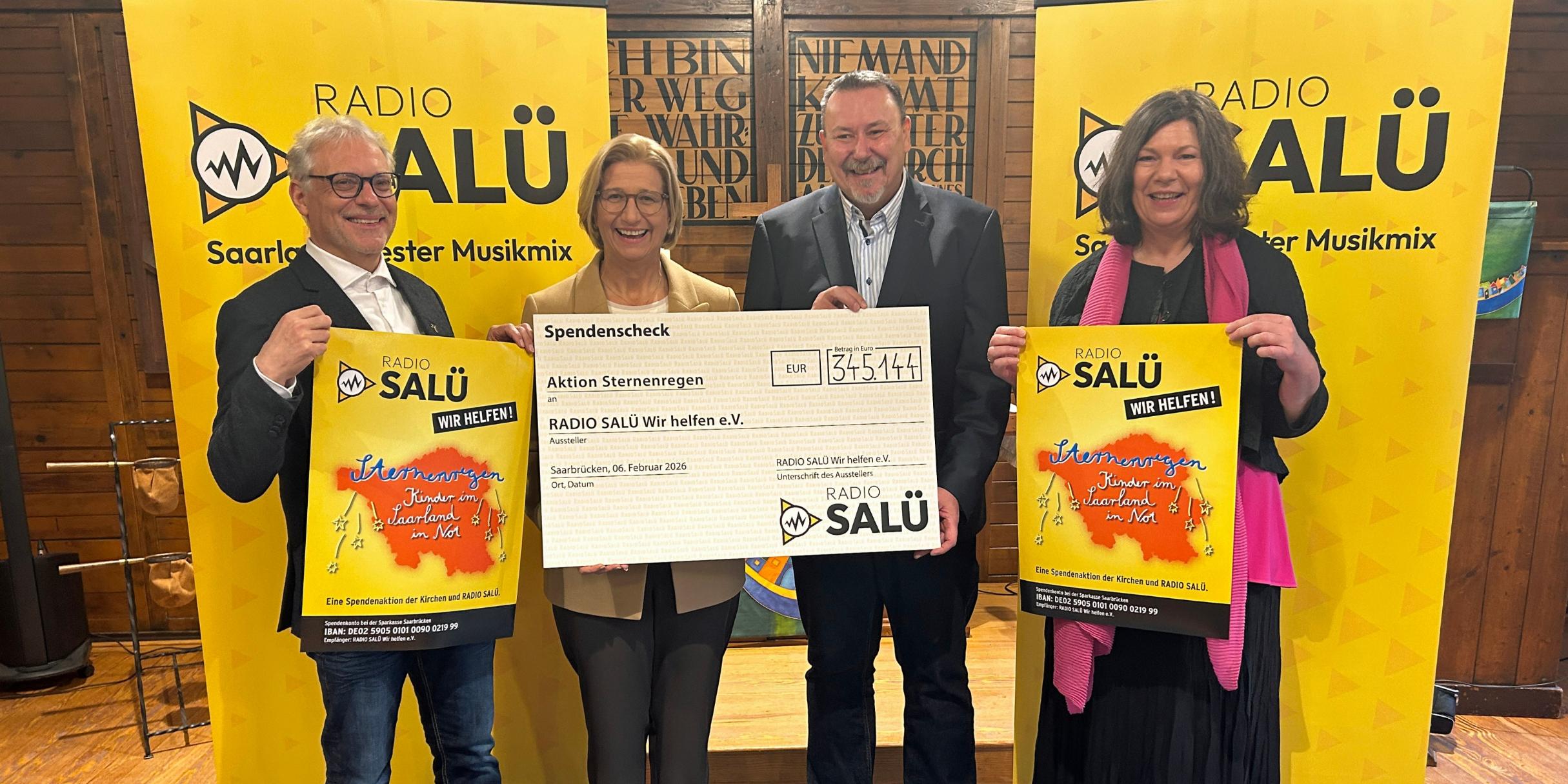 Vier Personen stehen in einem Innenraum vor zwei gelben Roll‑Up‑Bannern mit dem Logo von „Radio Salü“. Die beiden Personen in der Mitte halten gemeinsam einen großen Spendenscheck mit der Aufschrift „Aktion Sternenregen“ .Die beiden Personen außen halten jeweils ein farbiges Plakat derselben Spendenaktion. Der Boden besteht aus Holz, und im Hintergrund sind dunkle Holzwände mit eingravierten Schriftzügen zu sehen.