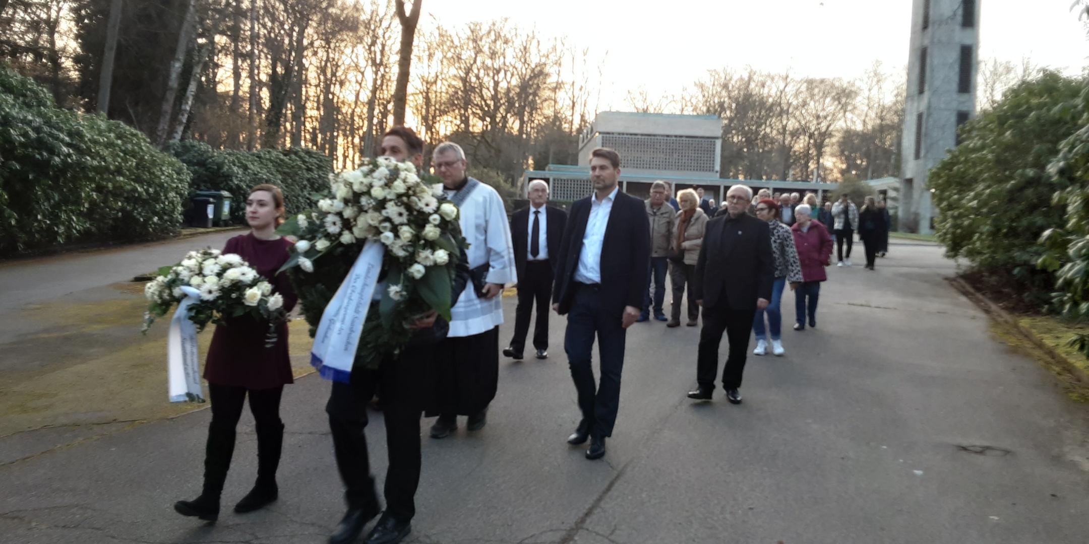 Eine Gruppe von Menschen geht in einem Trauerzug über einen breiten Friedhofsweg. Vorne tragen zwei Personen große weiße Kränze mit Trauerschleifen, begleitet von einer Person in liturgischer Kleidung. Dahinter folgen weitere Teilnehmende in respektvollem Abstand. Rechts im Hintergrund steht ein hoher Glockenturm, links und rechts säumen dichte Sträucher den Weg. Die Abendsonne scheint durch die kahlen Bäume und taucht die Szene in warmes Licht.