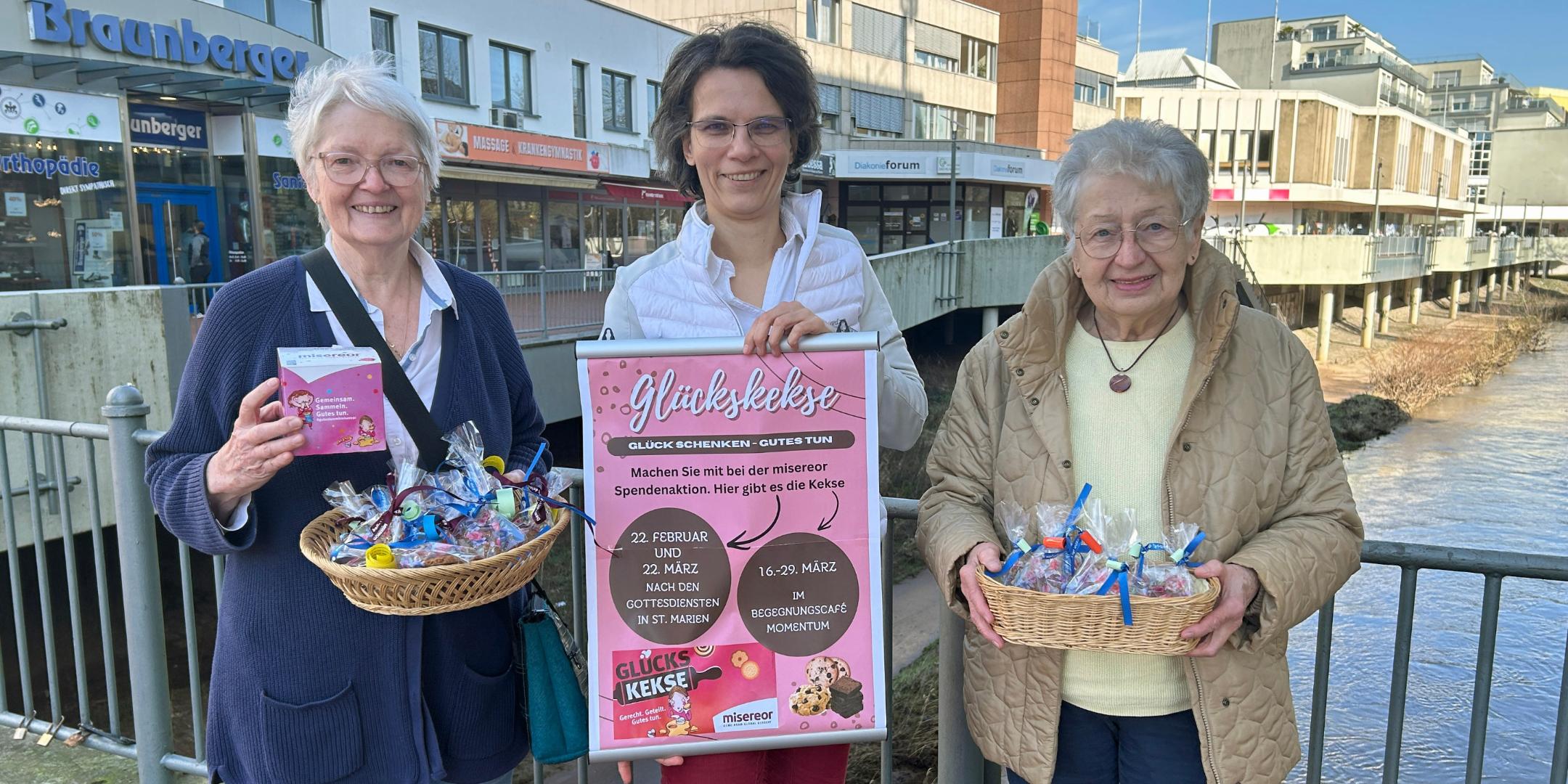 Drei Frauen stehen auf einer Brücke in der Neunkircher Innenstadt. Zwei von ihnen halten je einen Korb mit bunt verpackten Glückskeksen. Die Frau in der Mitte hält ein großes, pinkfarbenes Plakat mit Informationen zu einer Glückskeks‑Aktion. Im Hintergrund sind Geschäfte, Gebäude und die Blies zu sehen.