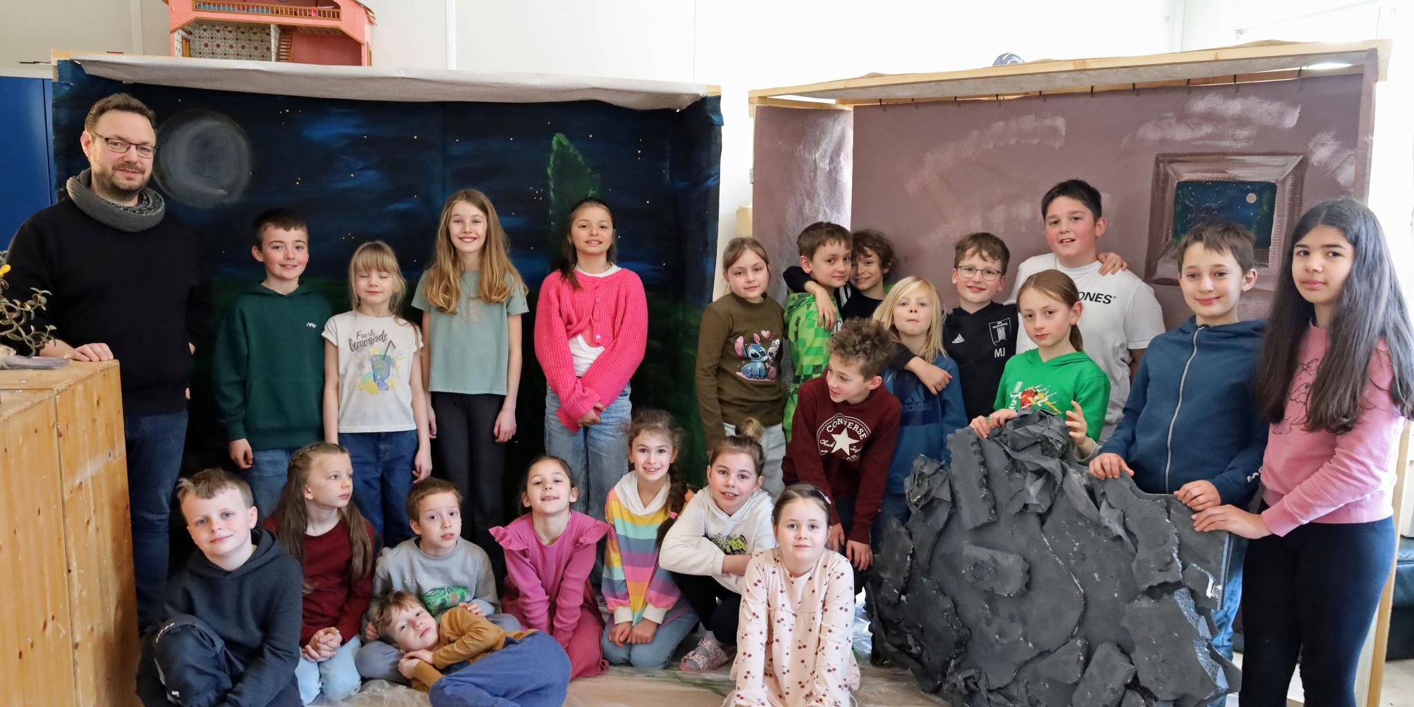 Eine größere Gruppe von Kindern und einer erwachsenen Person steht und sitzt in einem Raum zwischen zwei gebauten Kulissen. Links befindet sich eine Kulisse, die eine nächtliche Landschaft mit Mond und Sternen zeigt. Rechts steht eine zweite Kulisse in warmen Farben mit einem gemalten Fenster. Davor lehnt ein großes künstliches Felsstück. Einige Kinder sitzen auf dem Boden, andere stehen dahinter. Die Gruppe ist umgeben von Holz‑ und Bastelmaterialien, was auf ein gemeinsames kreatives Projekt oder eine Aufführung hindeutet.