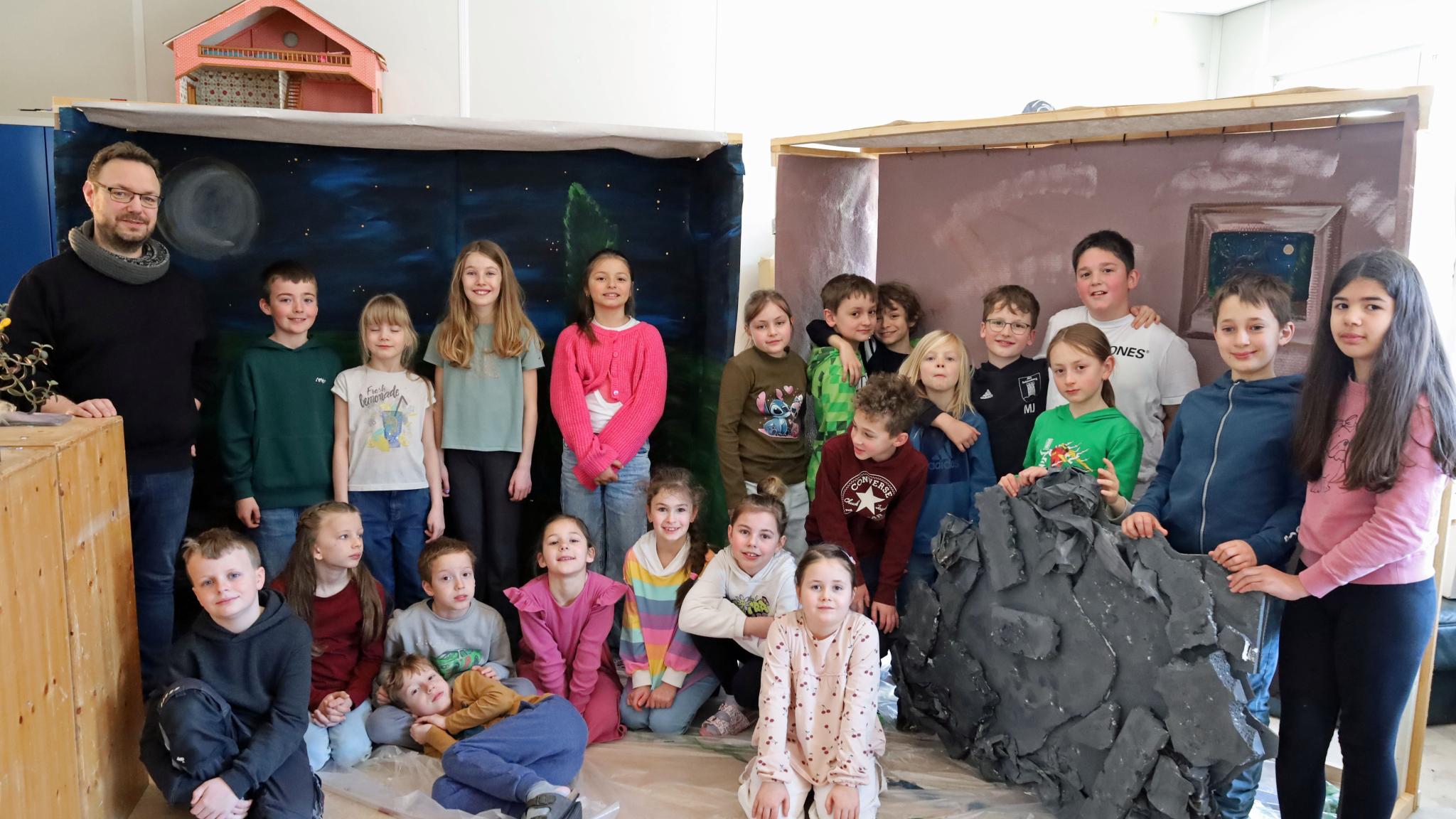 Eine größere Gruppe von Kindern und einer erwachsenen Person steht und sitzt in einem Raum zwischen zwei gebauten Kulissen. Links befindet sich eine Kulisse, die eine nächtliche Landschaft mit Mond und Sternen zeigt. Rechts steht eine zweite Kulisse in warmen Farben mit einem gemalten Fenster. Davor lehnt ein großes künstliches Felsstück. Einige Kinder sitzen auf dem Boden, andere stehen dahinter. Die Gruppe ist umgeben von Holz‑ und Bastelmaterialien, was auf ein gemeinsames kreatives Projekt oder eine Aufführung hindeutet.
