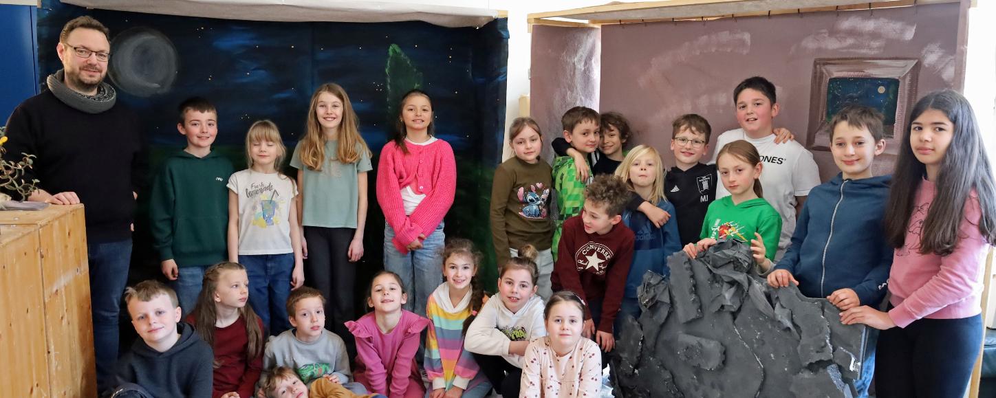 Eine größere Gruppe von Kindern und einer erwachsenen Person steht und sitzt in einem Raum zwischen zwei gebauten Kulissen. Links befindet sich eine Kulisse, die eine nächtliche Landschaft mit Mond und Sternen zeigt. Rechts steht eine zweite Kulisse in warmen Farben mit einem gemalten Fenster. Davor lehnt ein großes künstliches Felsstück. Einige Kinder sitzen auf dem Boden, andere stehen dahinter. Die Gruppe ist umgeben von Holz‑ und Bastelmaterialien, was auf ein gemeinsames kreatives Projekt oder eine Aufführung hindeutet.