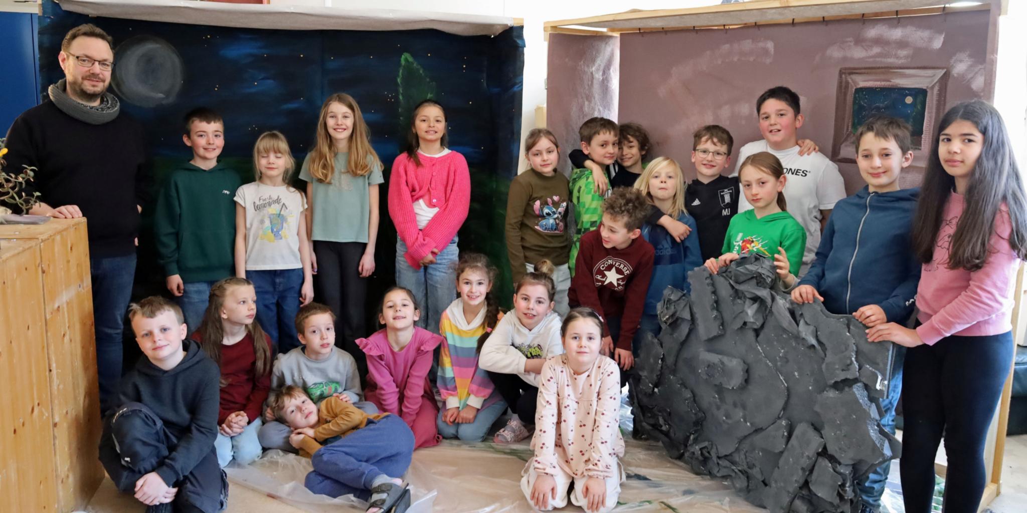Eine größere Gruppe von Kindern und einer erwachsenen Person steht und sitzt in einem Raum zwischen zwei gebauten Kulissen. Links befindet sich eine Kulisse, die eine nächtliche Landschaft mit Mond und Sternen zeigt. Rechts steht eine zweite Kulisse in warmen Farben mit einem gemalten Fenster. Davor lehnt ein großes künstliches Felsstück. Einige Kinder sitzen auf dem Boden, andere stehen dahinter. Die Gruppe ist umgeben von Holz‑ und Bastelmaterialien, was auf ein gemeinsames kreatives Projekt oder eine Aufführung hindeutet.