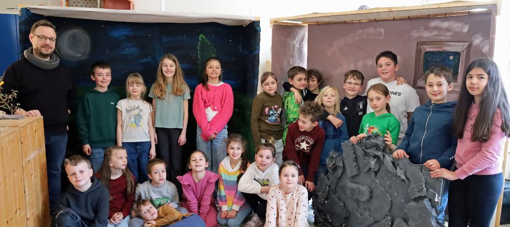 Eine größere Gruppe von Kindern und einer erwachsenen Person steht und sitzt in einem Raum zwischen zwei gebauten Kulissen. Links befindet sich eine Kulisse, die eine nächtliche Landschaft mit Mond und Sternen zeigt. Rechts steht eine zweite Kulisse in warmen Farben mit einem gemalten Fenster. Davor lehnt ein großes künstliches Felsstück. Einige Kinder sitzen auf dem Boden, andere stehen dahinter. Die Gruppe ist umgeben von Holz‑ und Bastelmaterialien, was auf ein gemeinsames kreatives Projekt oder eine Aufführung hindeutet.