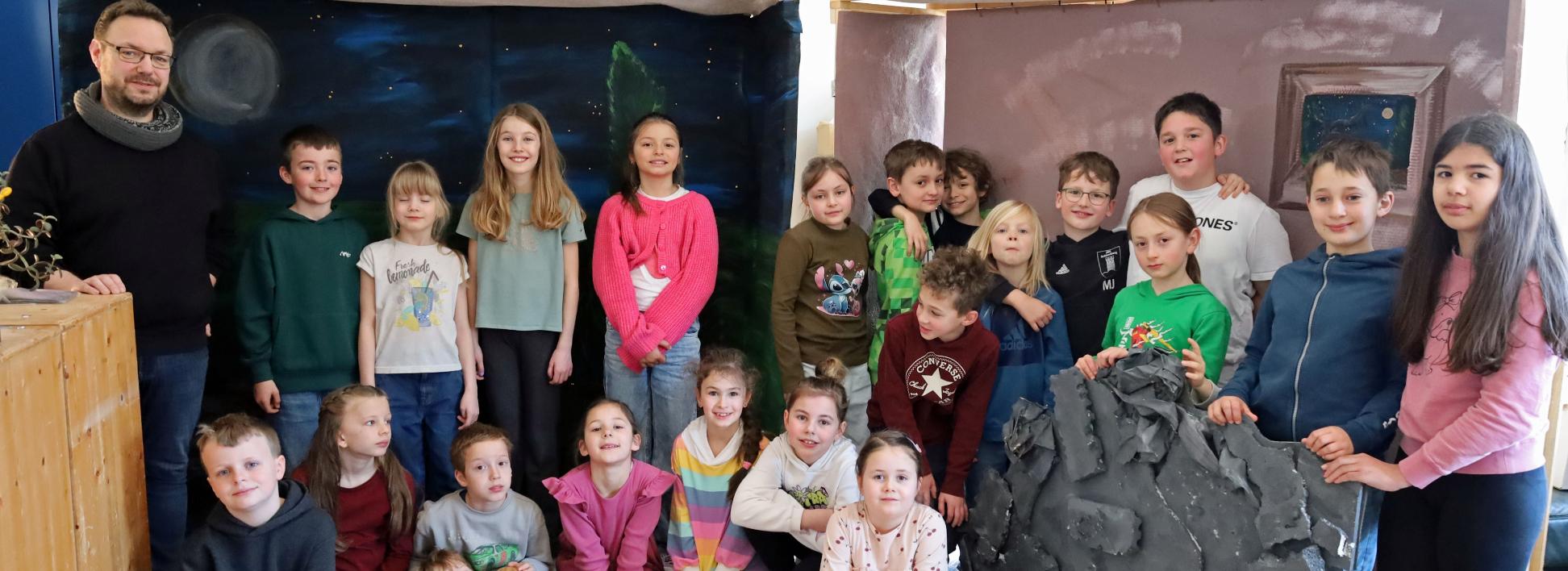 Eine größere Gruppe von Kindern und einer erwachsenen Person steht und sitzt in einem Raum zwischen zwei gebauten Kulissen. Links befindet sich eine Kulisse, die eine nächtliche Landschaft mit Mond und Sternen zeigt. Rechts steht eine zweite Kulisse in warmen Farben mit einem gemalten Fenster. Davor lehnt ein großes künstliches Felsstück. Einige Kinder sitzen auf dem Boden, andere stehen dahinter. Die Gruppe ist umgeben von Holz‑ und Bastelmaterialien, was auf ein gemeinsames kreatives Projekt oder eine Aufführung hindeutet.