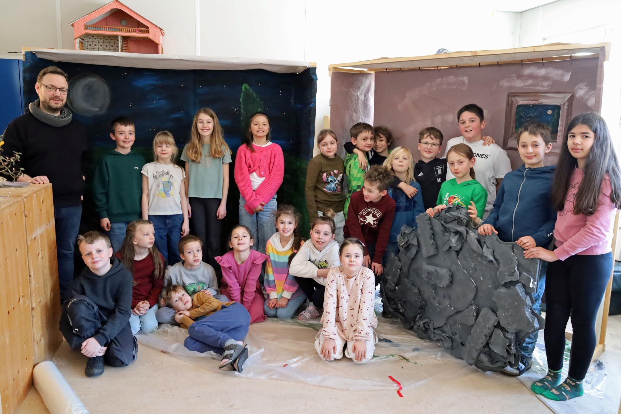 Eine größere Gruppe von Kindern und einer erwachsenen Person steht und sitzt in einem Raum zwischen zwei gebauten Kulissen. Links befindet sich eine Kulisse, die eine nächtliche Landschaft mit Mond und Sternen zeigt. Rechts steht eine zweite Kulisse in warmen Farben mit einem gemalten Fenster. Davor lehnt ein großes künstliches Felsstück. Einige Kinder sitzen auf dem Boden, andere stehen dahinter. Die Gruppe ist umgeben von Holz‑ und Bastelmaterialien, was auf ein gemeinsames kreatives Projekt oder eine Aufführung hindeutet.