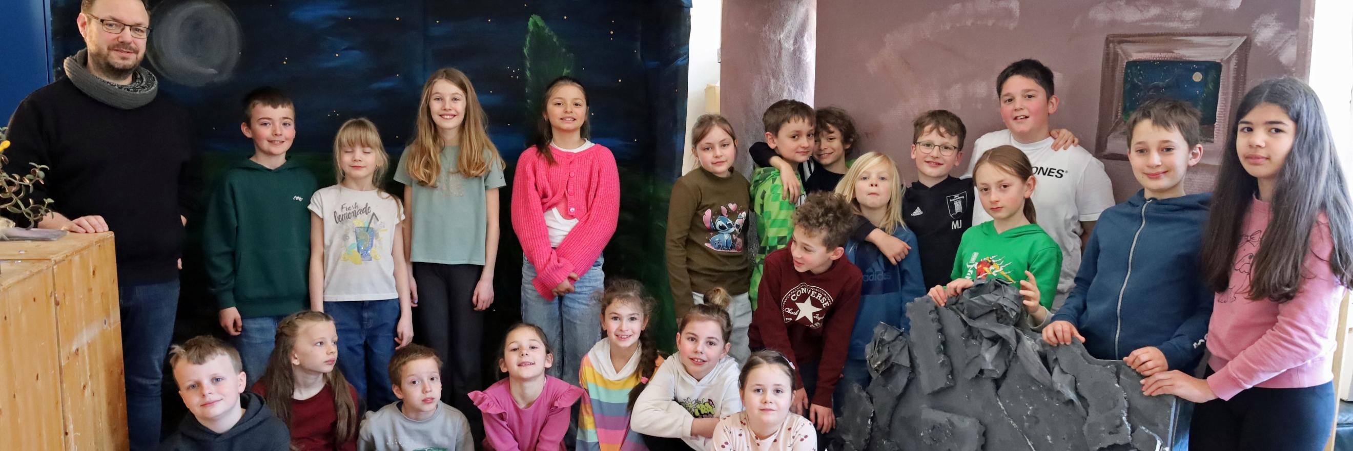 Eine größere Gruppe von Kindern und einer erwachsenen Person steht und sitzt in einem Raum zwischen zwei gebauten Kulissen. Links befindet sich eine Kulisse, die eine nächtliche Landschaft mit Mond und Sternen zeigt. Rechts steht eine zweite Kulisse in warmen Farben mit einem gemalten Fenster. Davor lehnt ein großes künstliches Felsstück. Einige Kinder sitzen auf dem Boden, andere stehen dahinter. Die Gruppe ist umgeben von Holz‑ und Bastelmaterialien, was auf ein gemeinsames kreatives Projekt oder eine Aufführung hindeutet.