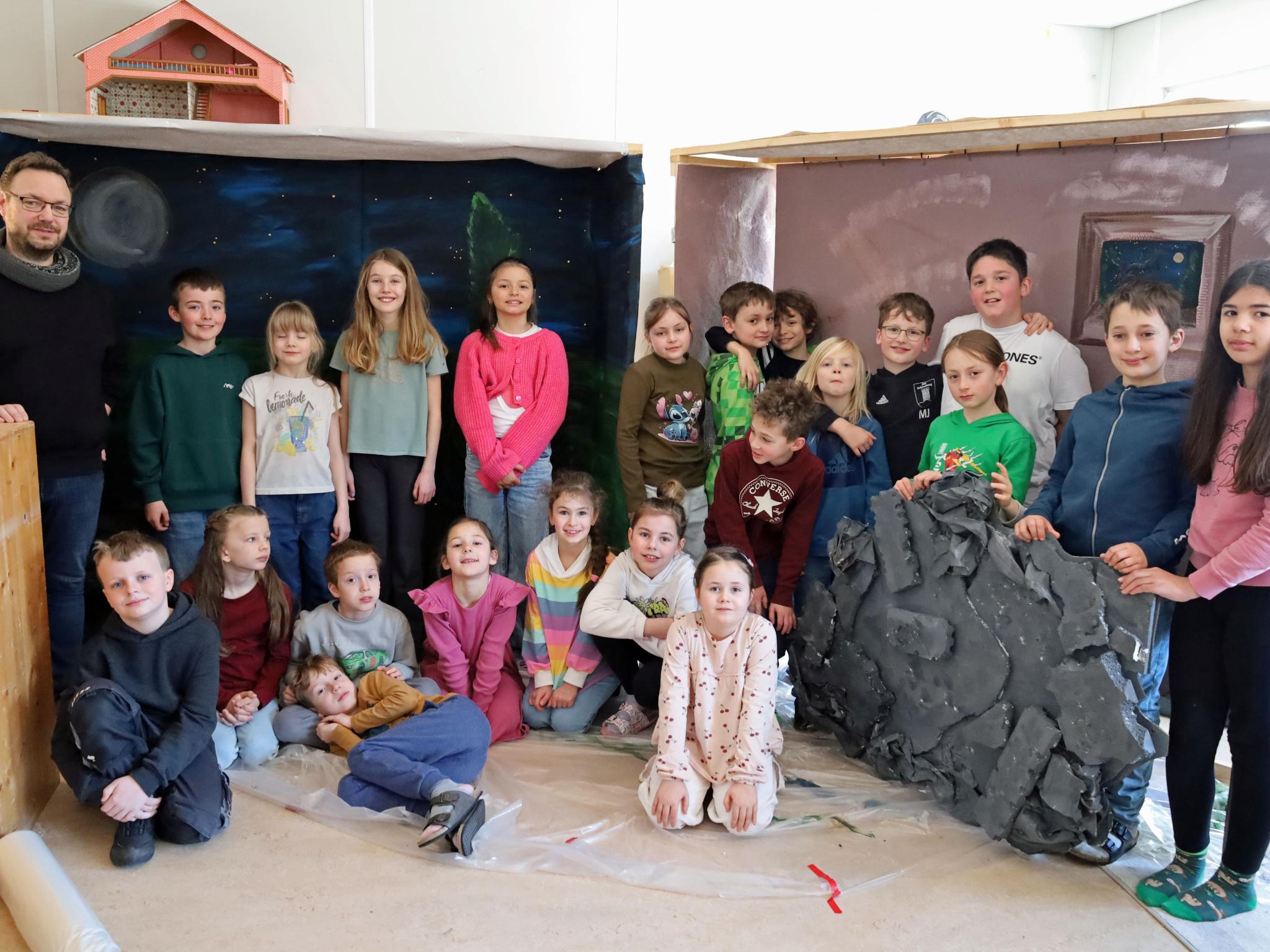 Eine größere Gruppe von Kindern und einer erwachsenen Person steht und sitzt in einem Raum zwischen zwei gebauten Kulissen. Links befindet sich eine Kulisse, die eine nächtliche Landschaft mit Mond und Sternen zeigt. Rechts steht eine zweite Kulisse in warmen Farben mit einem gemalten Fenster. Davor lehnt ein großes künstliches Felsstück. Einige Kinder sitzen auf dem Boden, andere stehen dahinter. Die Gruppe ist umgeben von Holz‑ und Bastelmaterialien, was auf ein gemeinsames kreatives Projekt oder eine Aufführung hindeutet.