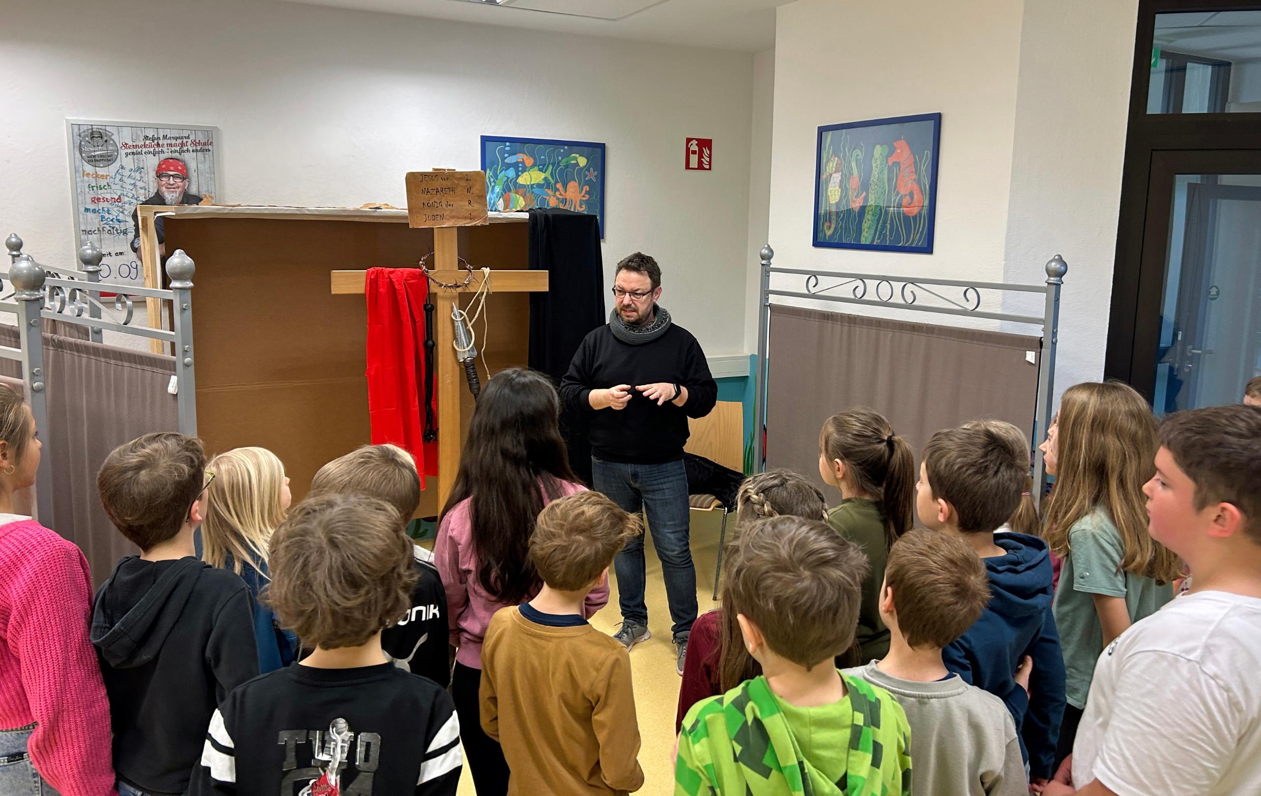 Eine größere Gruppe von Kindern und einer erwachsenen Person steht und sitzt in einem Raum zwischen zwei gebauten Kulissen. Links befindet sich eine Kulisse, die eine nächtliche Landschaft mit Mond und Sternen zeigt. Rechts steht eine zweite Kulisse in warmen Farben mit einem gemalten Fenster. Davor lehnt ein großes künstliches Felsstück. Einige Kinder sitzen auf dem Boden, andere stehen dahinter. Die Gruppe ist umgeben von Holz‑ und Bastelmaterialien, was auf ein gemeinsames kreatives Projekt oder eine Aufführung hindeutet.