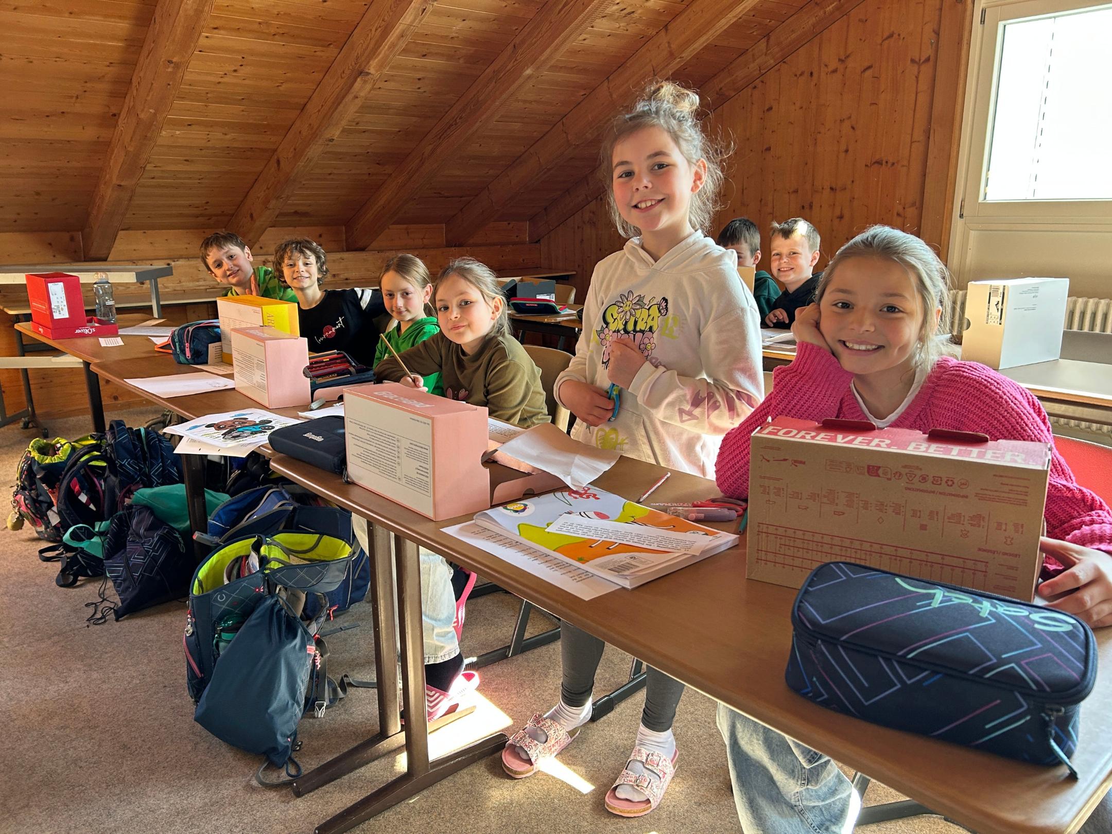 In einem Raum mit schräger Holzdecke sitzen mehrere Kinder an langen Tischen, die dicht nebeneinander stehen. Vor ihnen liegen Schulmaterialien, bunt gestaltete Blätter, Mappen und mehrere gebastelte Kartons, die wie kleine Projektkisten oder Modelle wirken. Auf dem Tisch stehen außerdem Federmäppchen und weitere Unterrichtsunterlagen. Ein Kind steht zwischen den Sitzplätzen und hält etwas in den Händen, während die übrigen Kinder sitzen und nach vorn schauen. Unter den Tischen stehen Schultaschen und Rucksäcke. Durch ein Fenster auf der rechten Seite fällt helles Tageslicht in den Raum. Die Szene wirkt wie eine Unterrichts- oder Projektpräsentation.