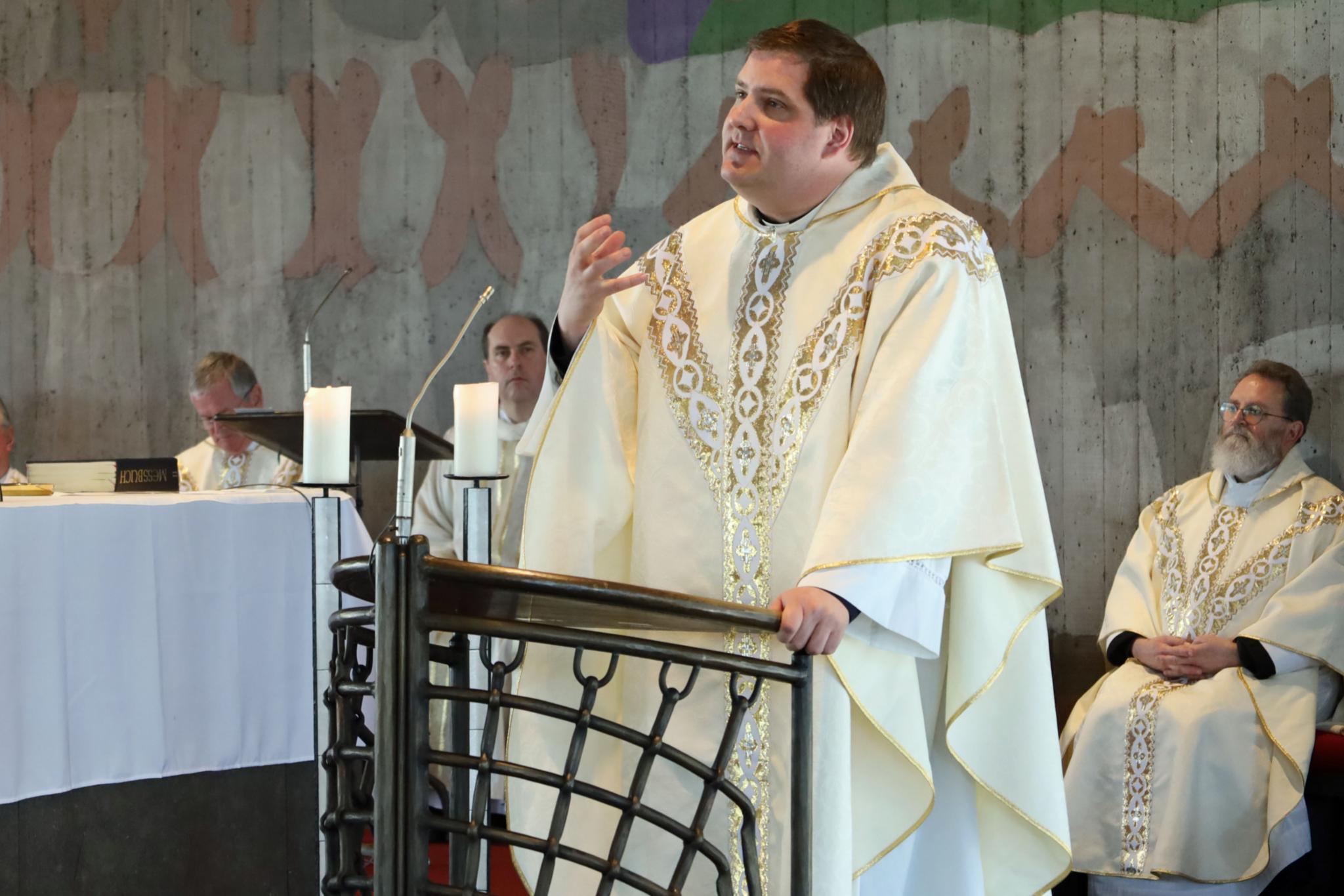 Ein Priester in einem reich verzierten, cremefarbenen liturgischen Gewand steht am Ambo einer Kirche und hält eine Predigt. Die Person hebt eine Hand leicht an, als würde ein Gedankengang betont. Links davon sitzt eine weitere Person in liturgischer Kleidung hinter einem mit weißem Tuch bedeckten Altar, auf dem Kerzen und Bücher liegen. Rechts im Bild sitzt einen weiteren Priester ebenfalls in festlichem Gewand. Im Hintergrund ist eine Wand mit farbigen, abstrakten Malereien zu sehen. Die Szene zeigt einen Moment während eines feierlichen Gottesdienstes.