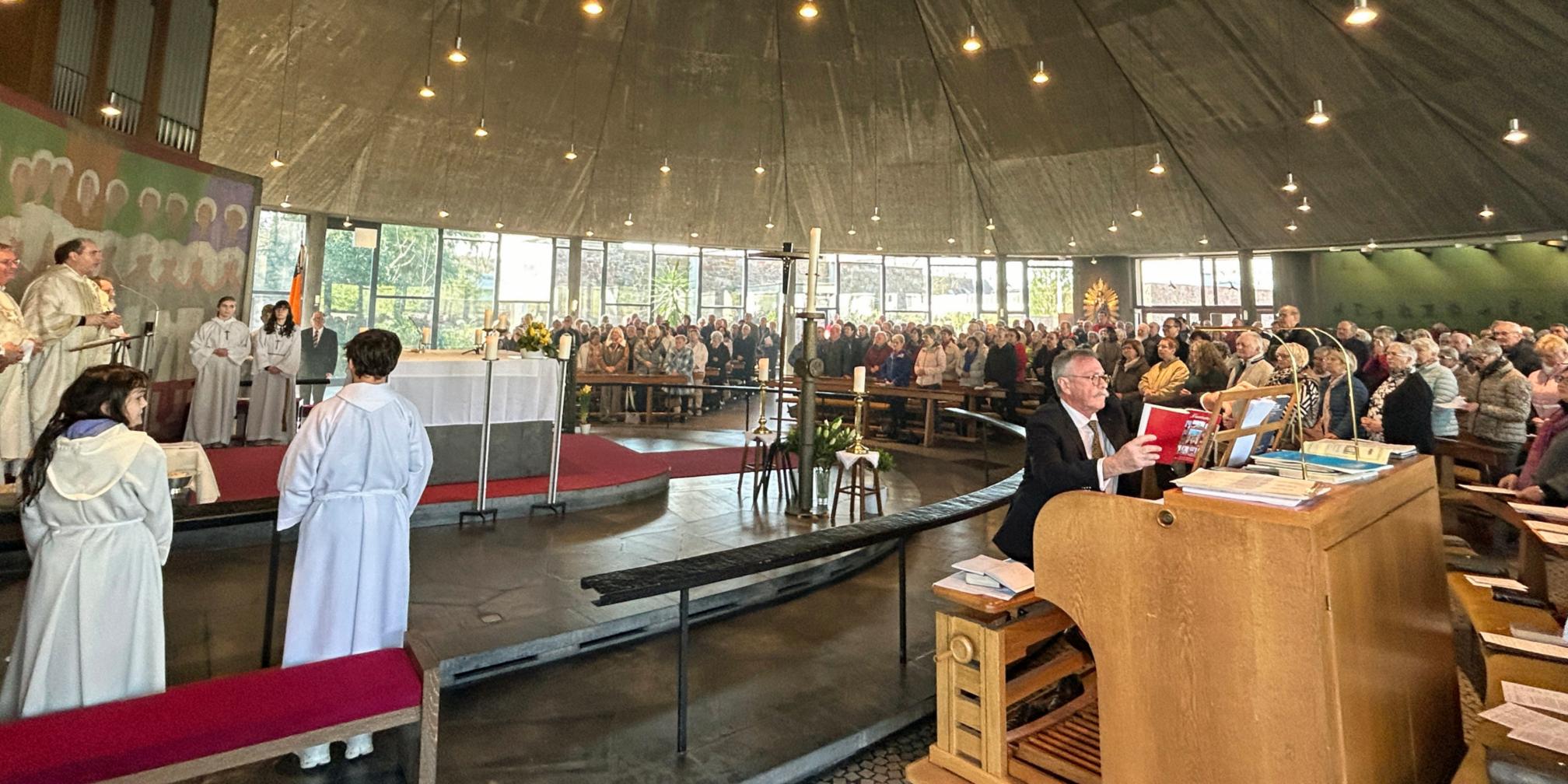 Innenaufnahme einer großen, modern gestalteten Kirche während eines festlichen Gottesdienstes. Die Gemeinde füllt den gesamten Raum, viele Personen sitzen oder stehen dicht beieinander in den Bankreihen, die halbkreisförmig um den Altar angeordnet sind. Der Altar ist zentral positioniert, weiß gedeckt und von liturgischen Diensten umgeben, darunter mehrere Personen in hellen liturgischen Gewändern. Im linken Bereich stehen weitere Personen im Altarraum und beteiligen sich am Gottesdienst. Rechts im Vordergrund sitzt eine Person an einer großen Orgel und spielt, vor sich Noten und Bücher auf dem Instrument. Der Kirchenraum ist lichtdurchflutet durch hohe Fenster im Hintergrund, und zahlreiche kleine Lampen hängen von der dunklen, zeltartigen Decke herab. Insgesamt vermittelt das Bild den Eindruck eines feierlichen und gut besuchten Festhochamts.