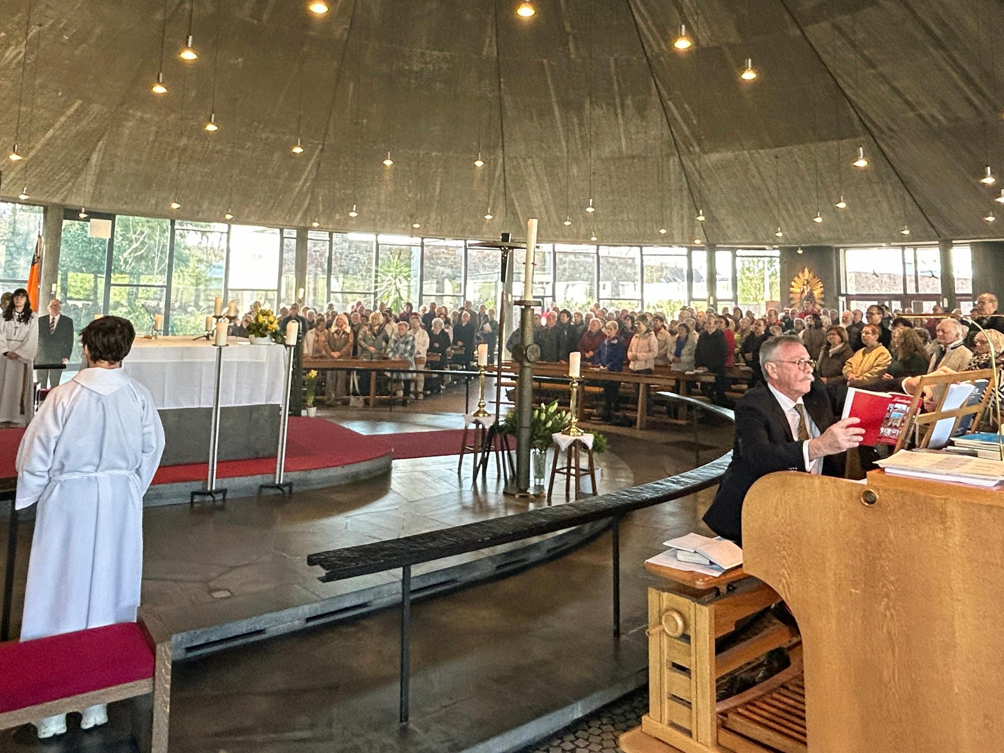 Innenaufnahme einer großen, modern gestalteten Kirche während eines festlichen Gottesdienstes. Die Gemeinde füllt den gesamten Raum, viele Personen sitzen oder stehen dicht beieinander in den Bankreihen, die halbkreisförmig um den Altar angeordnet sind. Der Altar ist zentral positioniert, weiß gedeckt und von liturgischen Diensten umgeben, darunter mehrere Personen in hellen liturgischen Gewändern. Im linken Bereich stehen weitere Personen im Altarraum und beteiligen sich am Gottesdienst. Rechts im Vordergrund sitzt eine Person an einer großen Orgel und spielt, vor sich Noten und Bücher auf dem Instrument. Der Kirchenraum ist lichtdurchflutet durch hohe Fenster im Hintergrund, und zahlreiche kleine Lampen hängen von der dunklen, zeltartigen Decke herab. Insgesamt vermittelt das Bild den Eindruck eines feierlichen und gut besuchten Festhochamts.