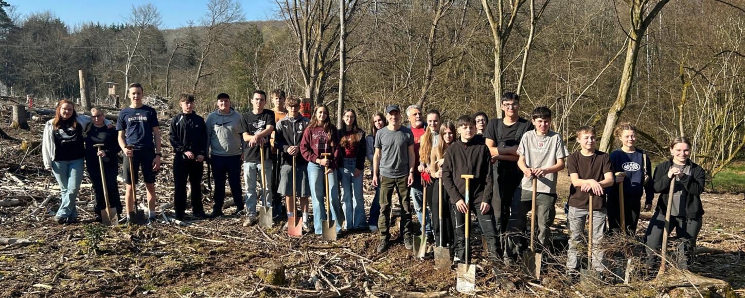 Eine große Gruppe Jugendlicher und Erwachsener steht auf einer offenen, kahlen Waldfläche zwischen dünnen, blattlosen Bäumen. Der Boden ist mit Ästen, Rindenschollen und Holzresten bedeckt. Viele der Teilnehmenden halten Spaten in den Händen und stehen in einer Reihe für ein Gruppenfoto. Im Hintergrund sind mehr Bäume, sanfte Hügel und ein blauer Himmel mit etwas Sonnenschein zu sehen.