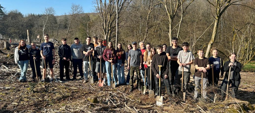 Eine große Gruppe Jugendlicher und Erwachsener steht auf einer offenen, kahlen Waldfläche zwischen dünnen, blattlosen Bäumen. Der Boden ist mit Ästen, Rindenschollen und Holzresten bedeckt. Viele der Teilnehmenden halten Spaten in den Händen und stehen in einer Reihe für ein Gruppenfoto. Im Hintergrund sind mehr Bäume, sanfte Hügel und ein blauer Himmel mit etwas Sonnenschein zu sehen.
