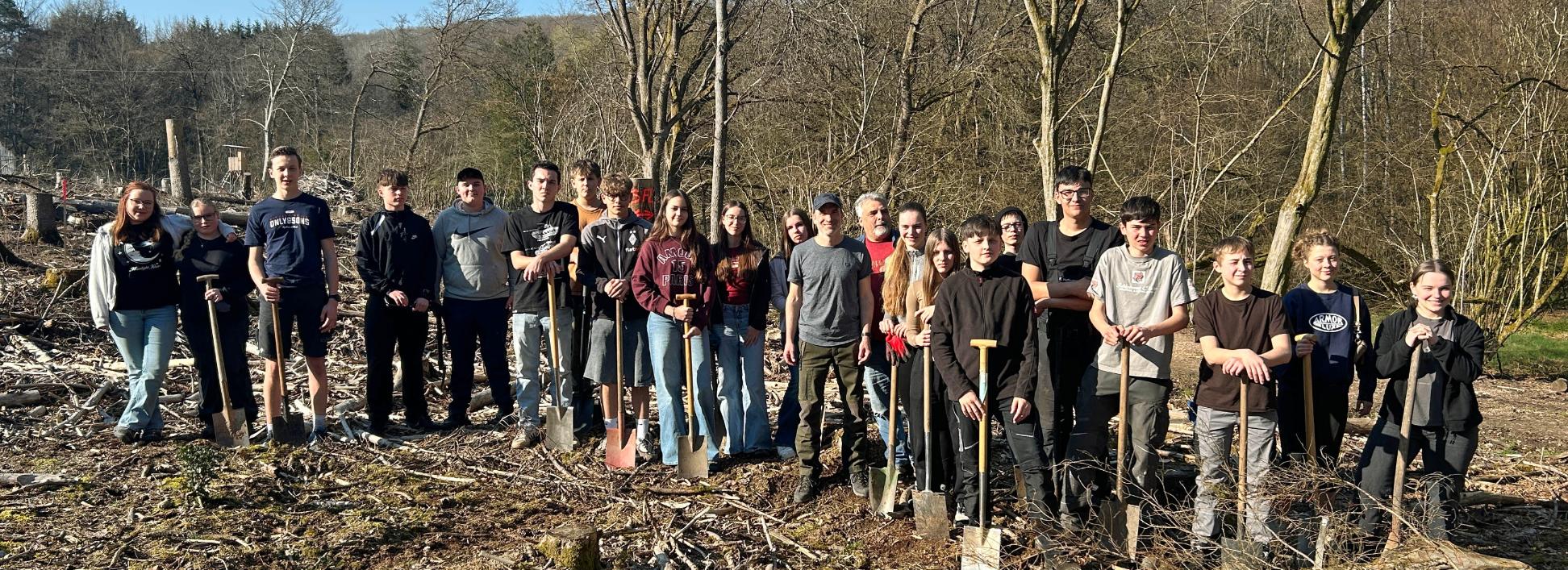 Eine große Gruppe Jugendlicher und Erwachsener steht auf einer offenen, kahlen Waldfläche zwischen dünnen, blattlosen Bäumen. Der Boden ist mit Ästen, Rindenschollen und Holzresten bedeckt. Viele der Teilnehmenden halten Spaten in den Händen und stehen in einer Reihe für ein Gruppenfoto. Im Hintergrund sind mehr Bäume, sanfte Hügel und ein blauer Himmel mit etwas Sonnenschein zu sehen.
